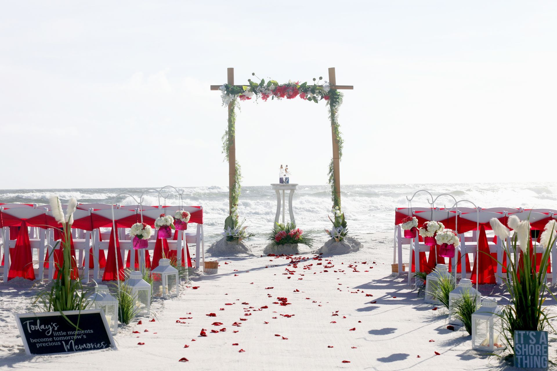 Beach wedding ceremony setup with red chairs, floral arch, and rose petals on white sand