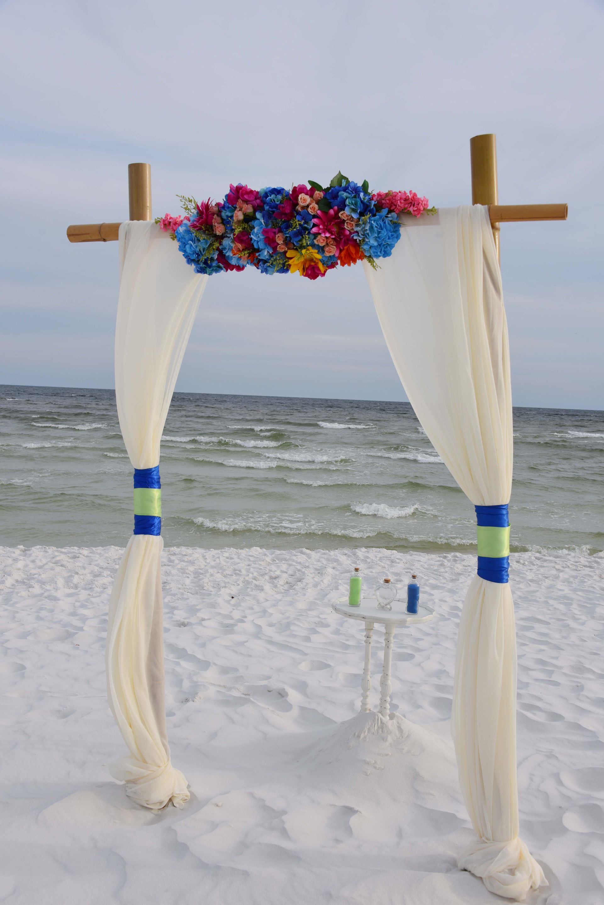 Beach wedding arch with white drapes, colorful flowers, and the ocean behind it.