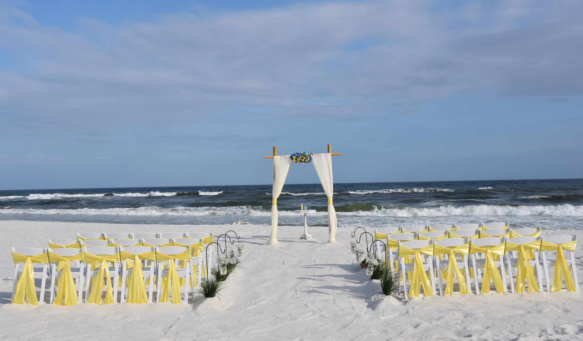 Beach wedding setup with white arch and rows of yellow chairs on a sandy shore under a cloudy sky