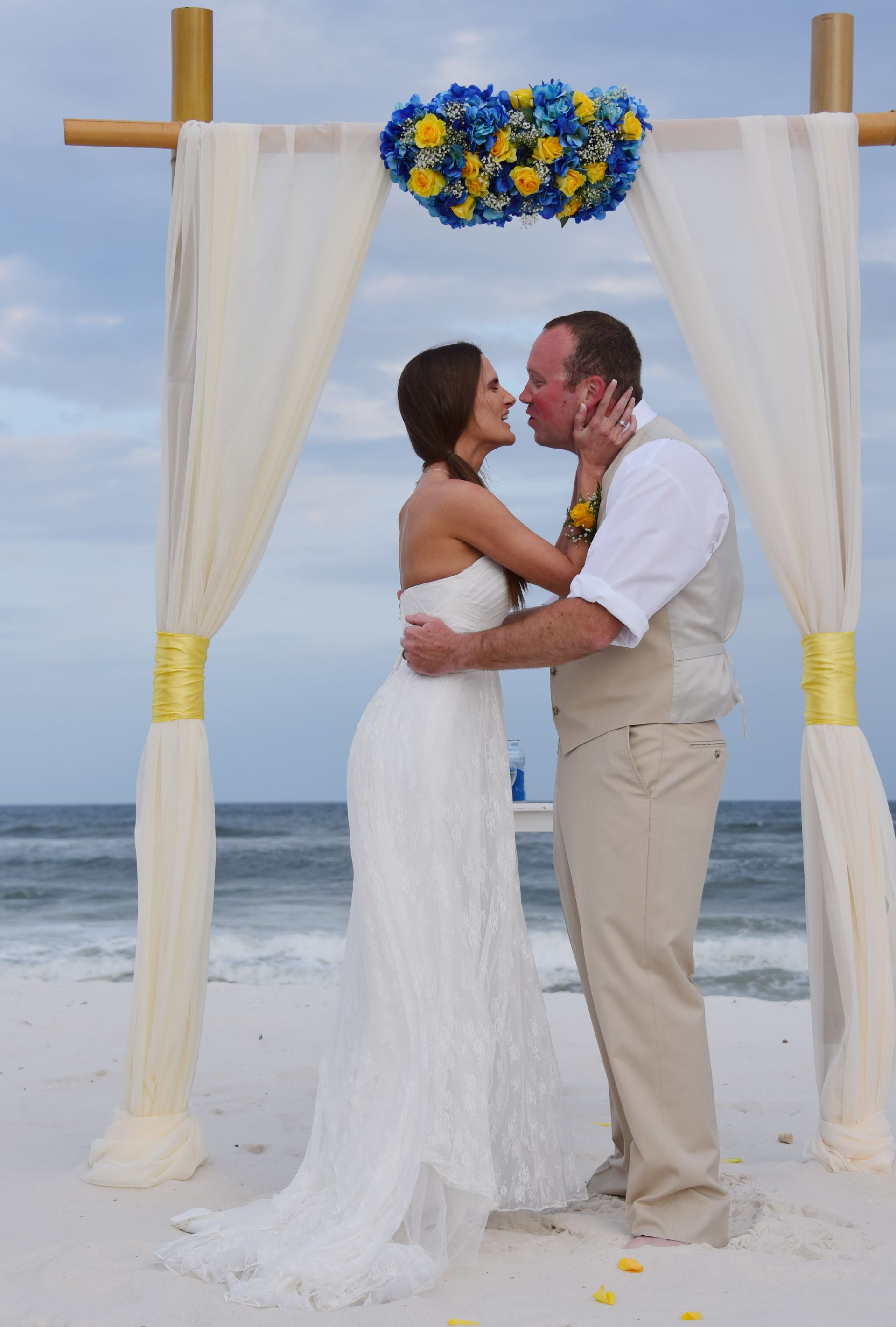 Couple embracing at a beach wedding arch with white drapes and blue flowers.