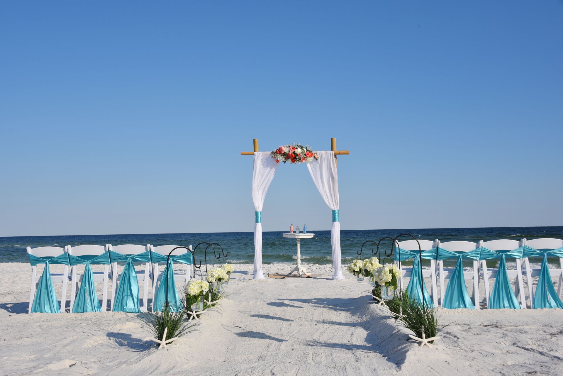 Beach wedding arch with white drapes and turquoise chairs on a sandy shore under a clear blue sky