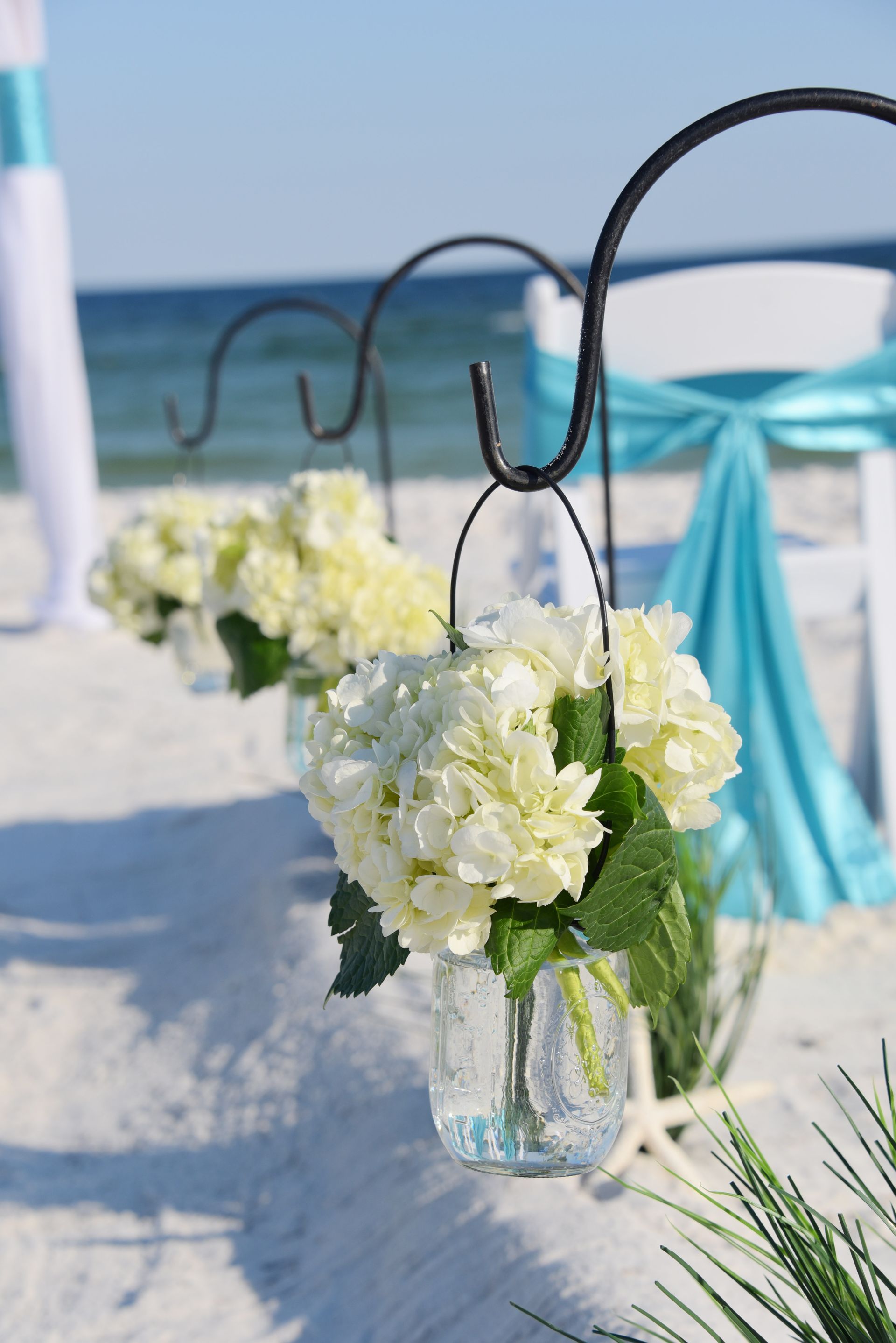 White flower bouquets hang from black hooks along a beach wedding aisle by the ocean.