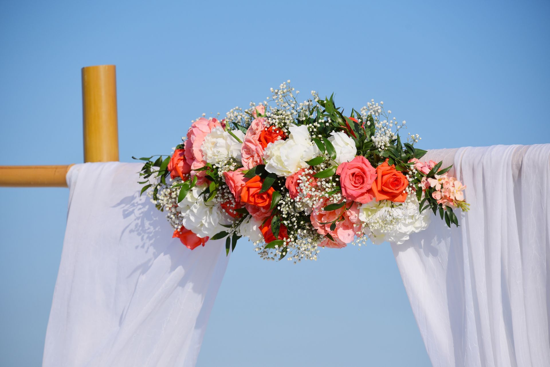 Floral arrangement of pink and white roses draped over a white fabric arch against a blue sky
