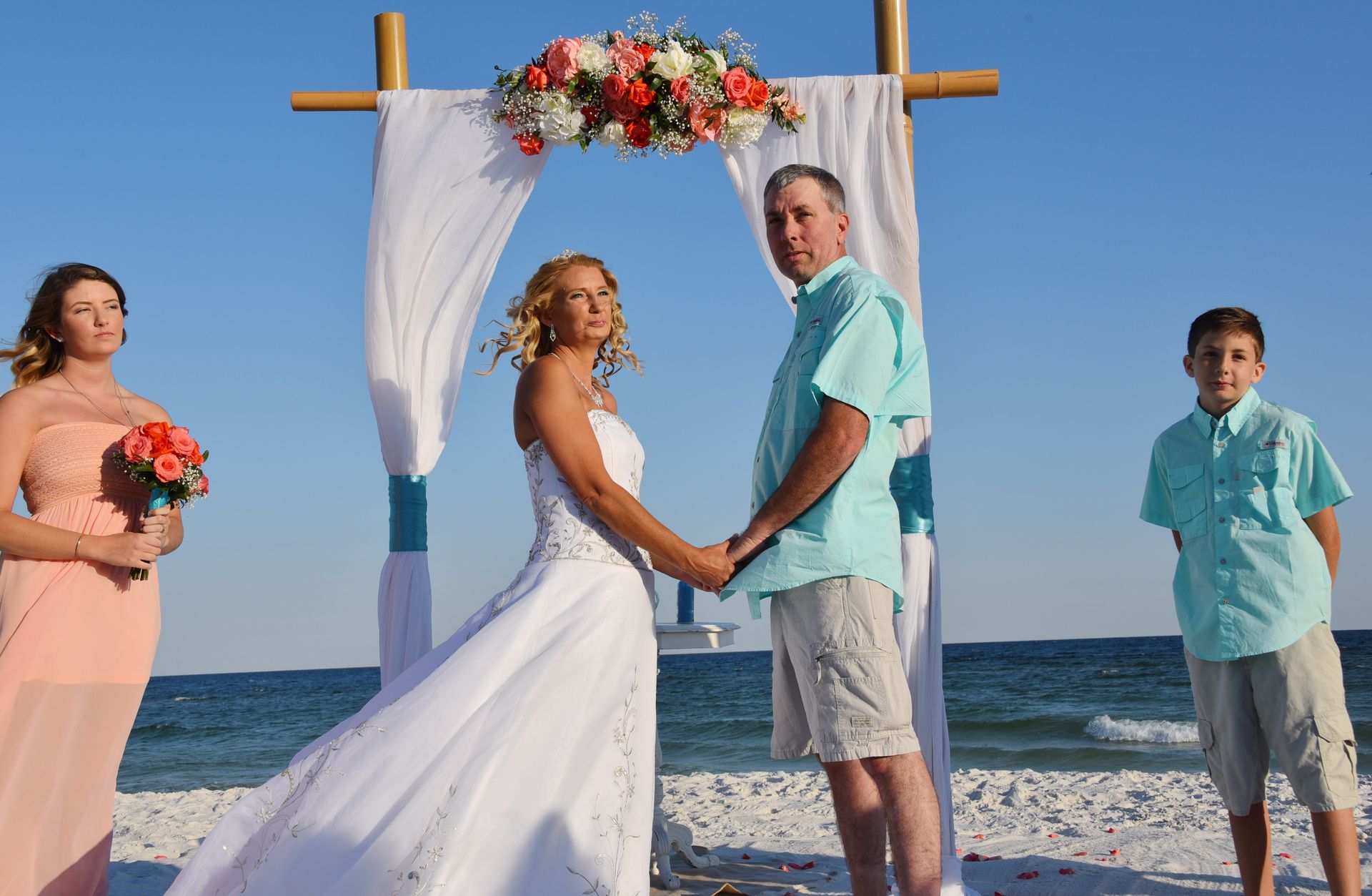 Beach wedding ceremony under a floral arch, bride and groom holding hands, bridesmaid and boy standing nearby.