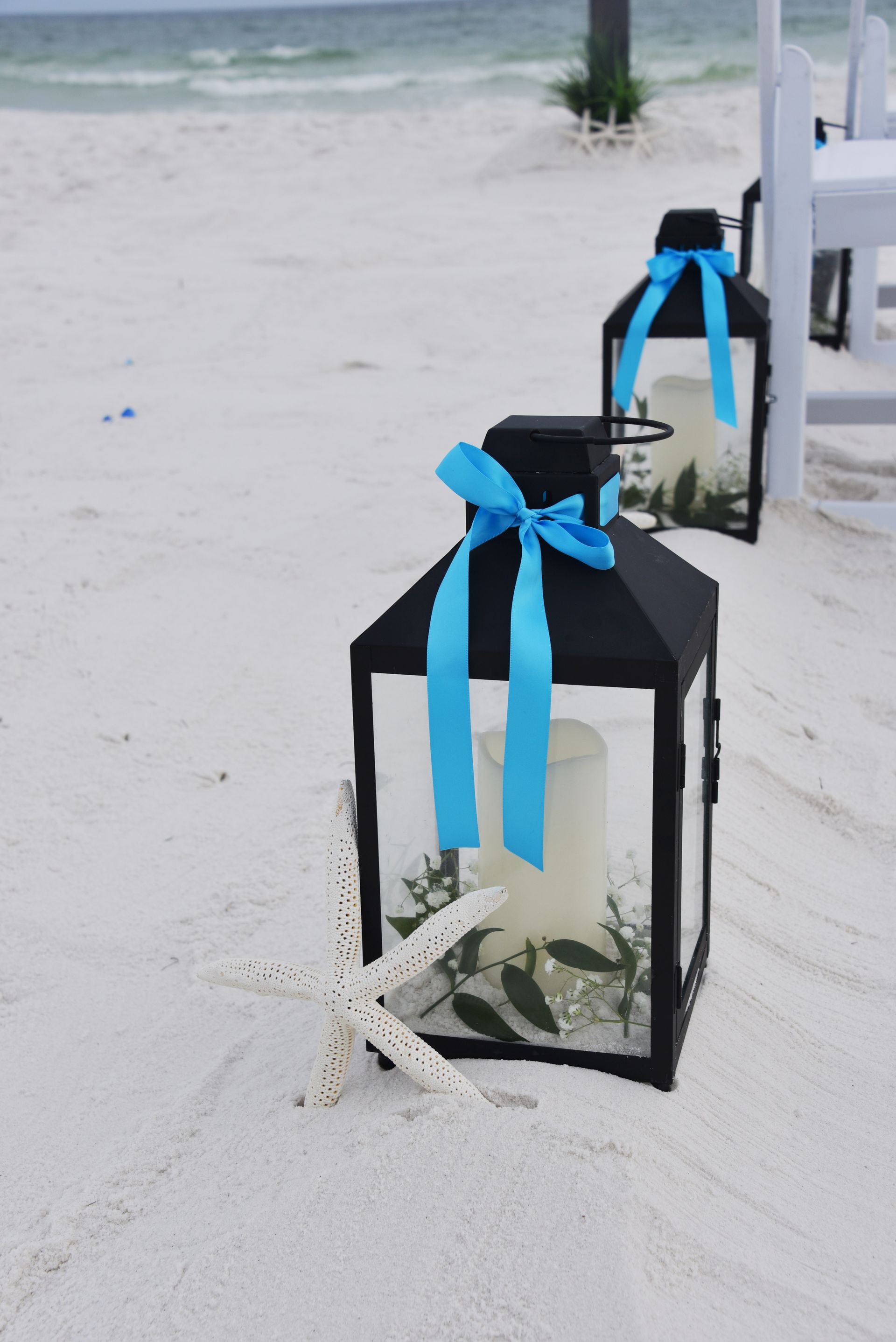 Black lantern with blue ribbon and white flowers on sandy beach, with more lanterns behind it