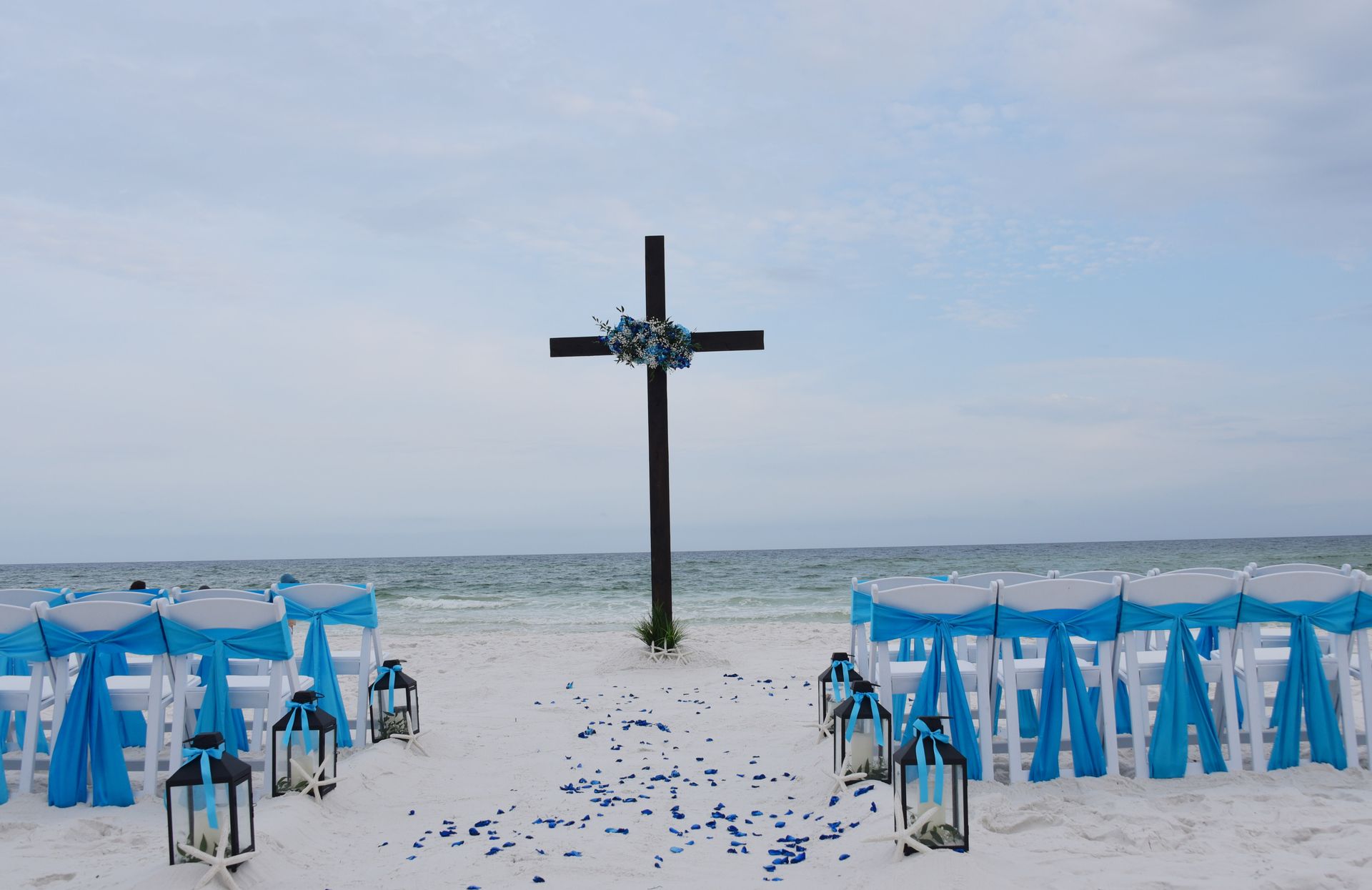 Beach wedding setup with blue chairs and lanterns facing a decorated cross by the sea