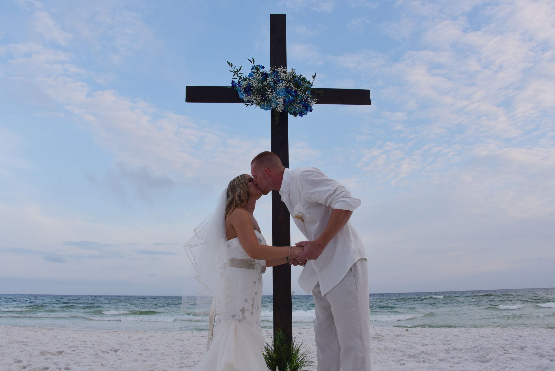 Couple in white kissing beneath a cross on a beach, with blue flowers against the ocean backdrop