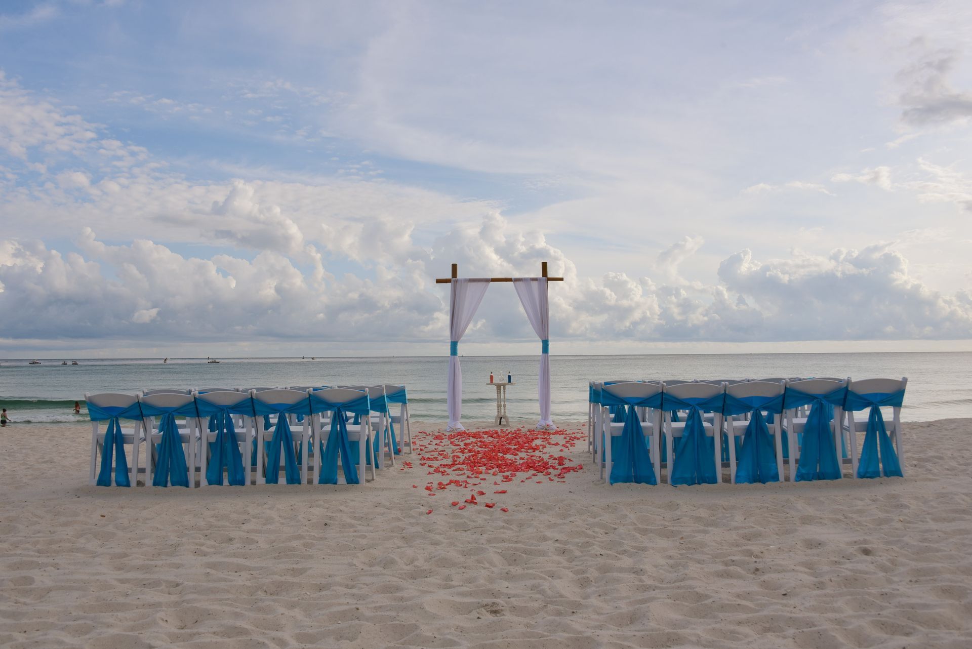 Beach wedding setup with blue chairs and a white arch, red petals scattered on the sand, ocean behind.