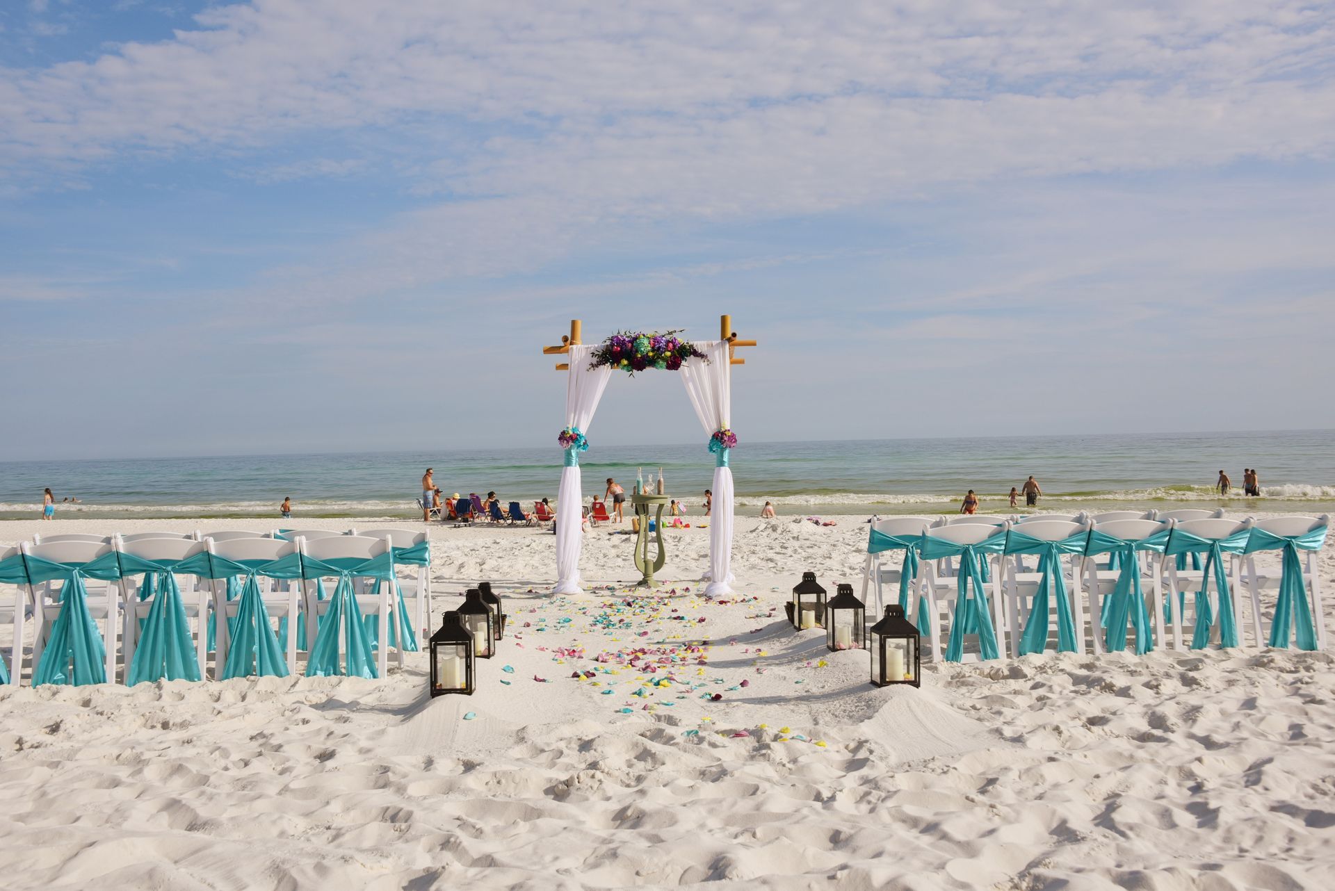Beach wedding setup with white arch, turquoise chairs, lanterns, and flowers on the sand by the ocean