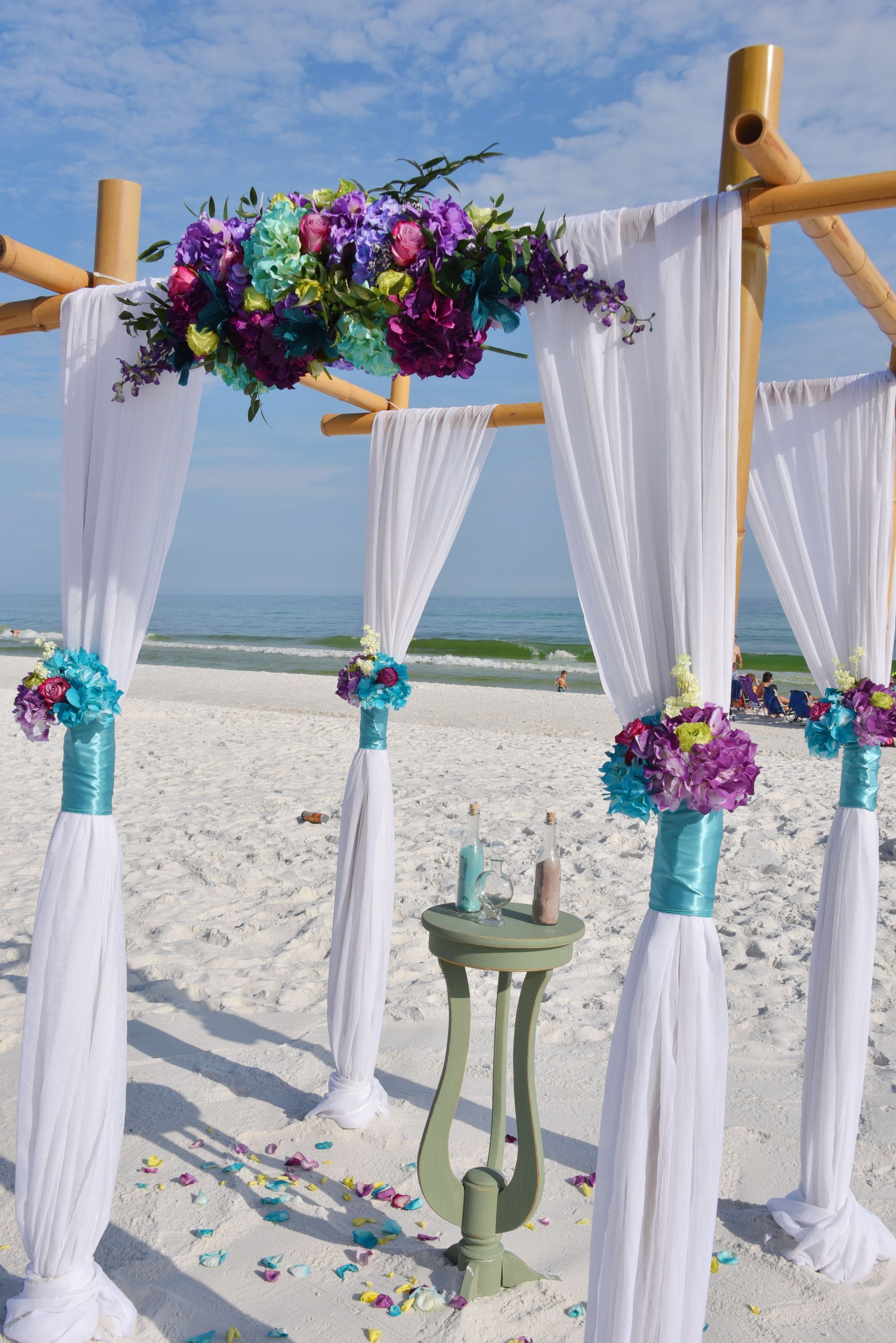 Beach wedding arch with white drapes, colorful flowers, and a small table on the sand near the ocean