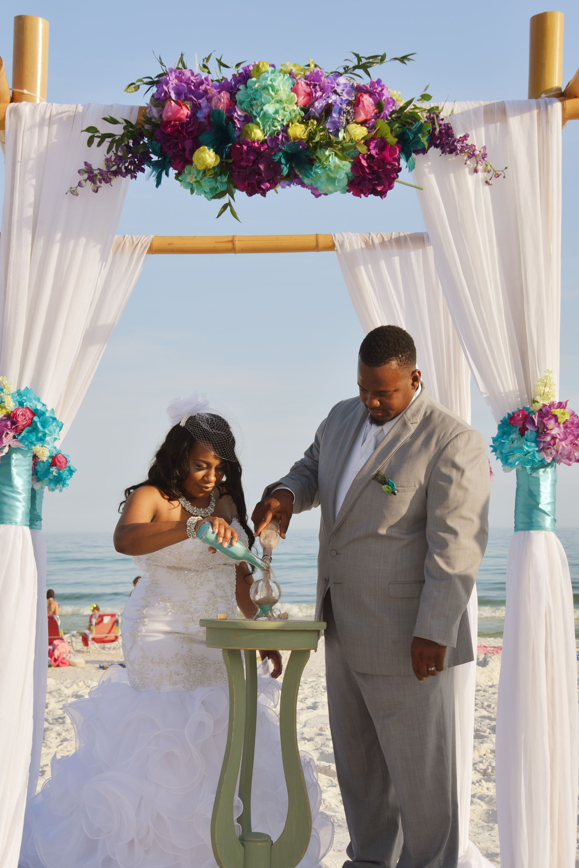 Couple signing a wedding document under a floral arch on a beach, with white drapes and turquoise ocean behind.