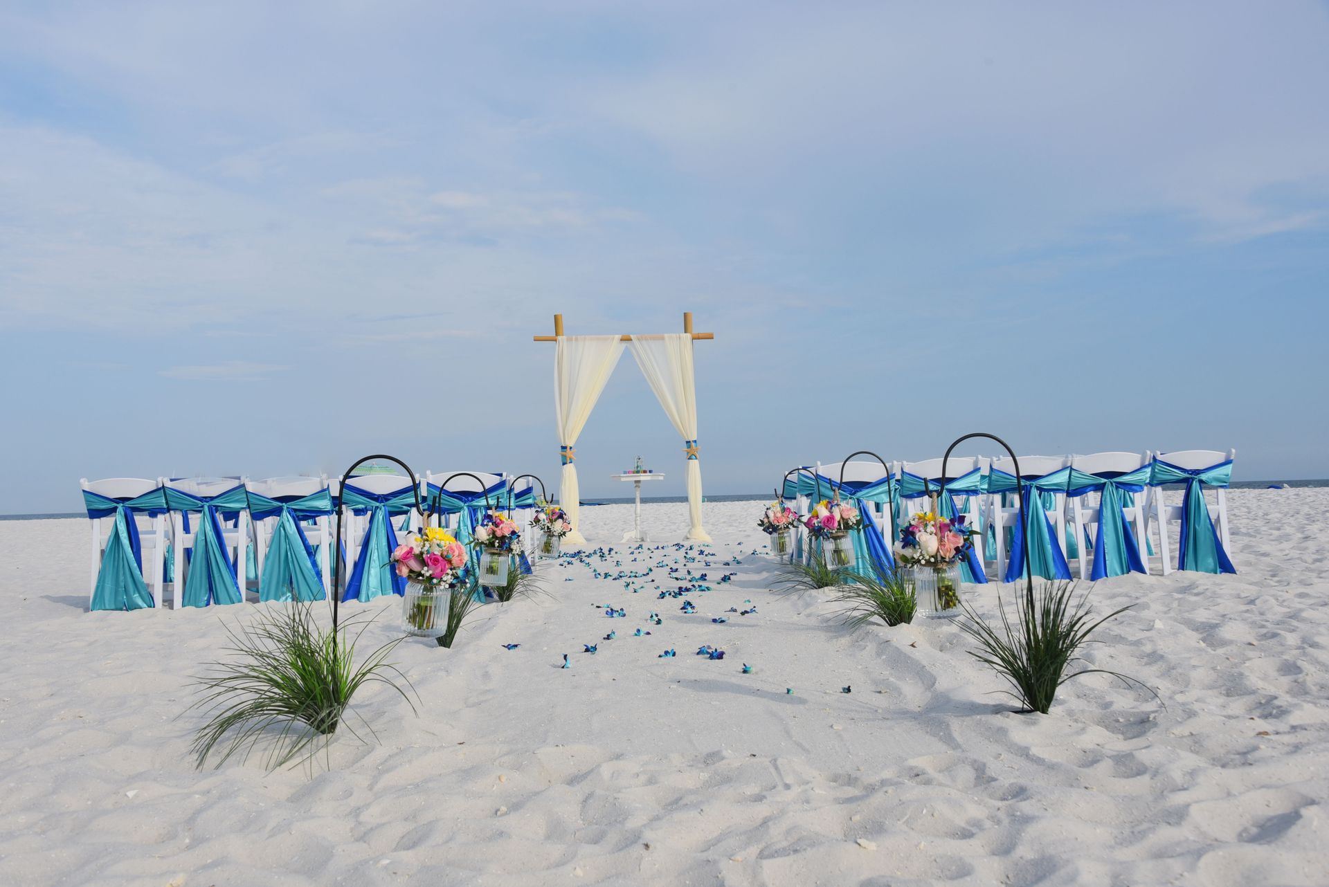 Beach wedding setup with white drapes, blue chairs, and flower-lined aisle on white sand