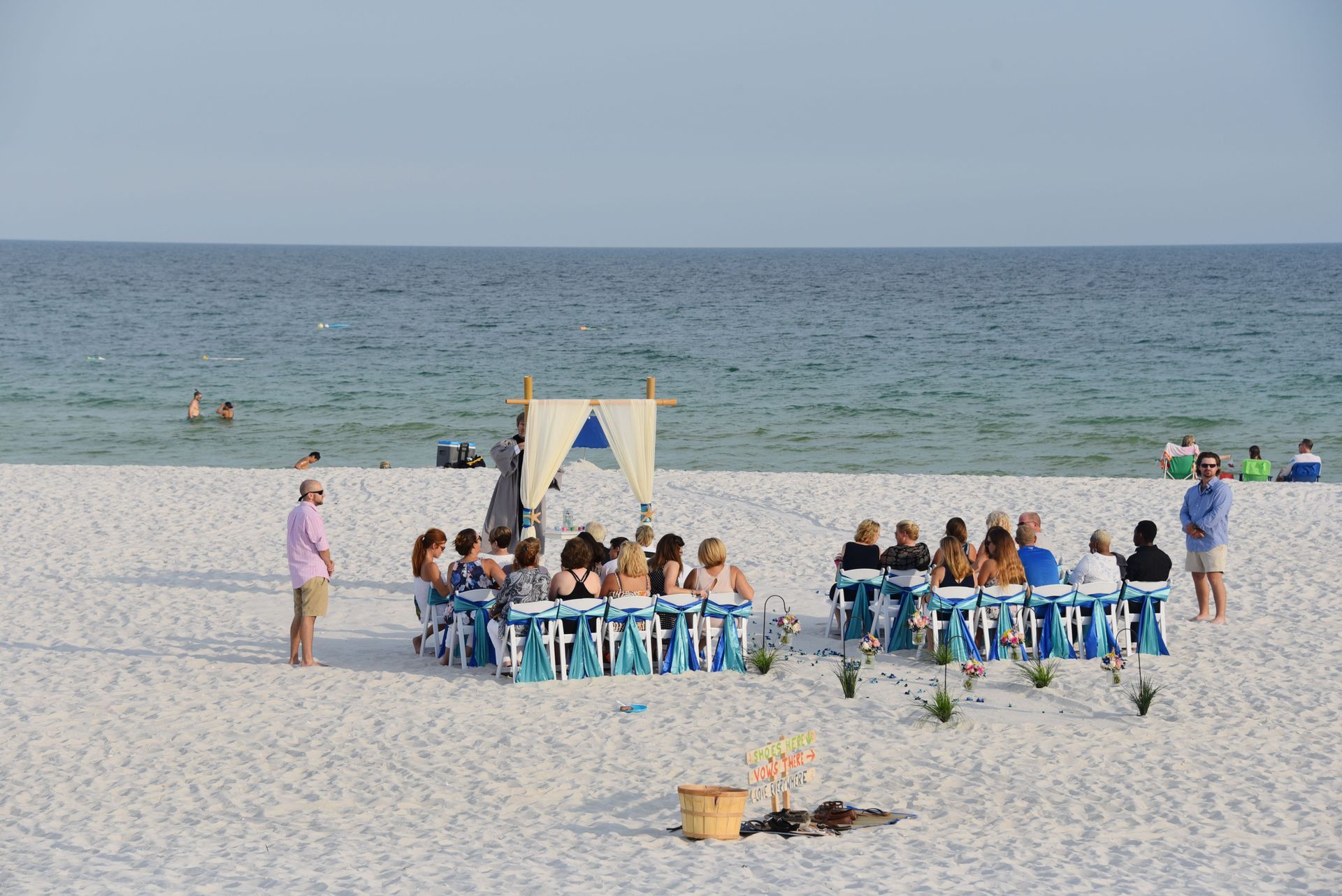 Group seated at blue tables on a sandy beach beside the ocean under a canopy