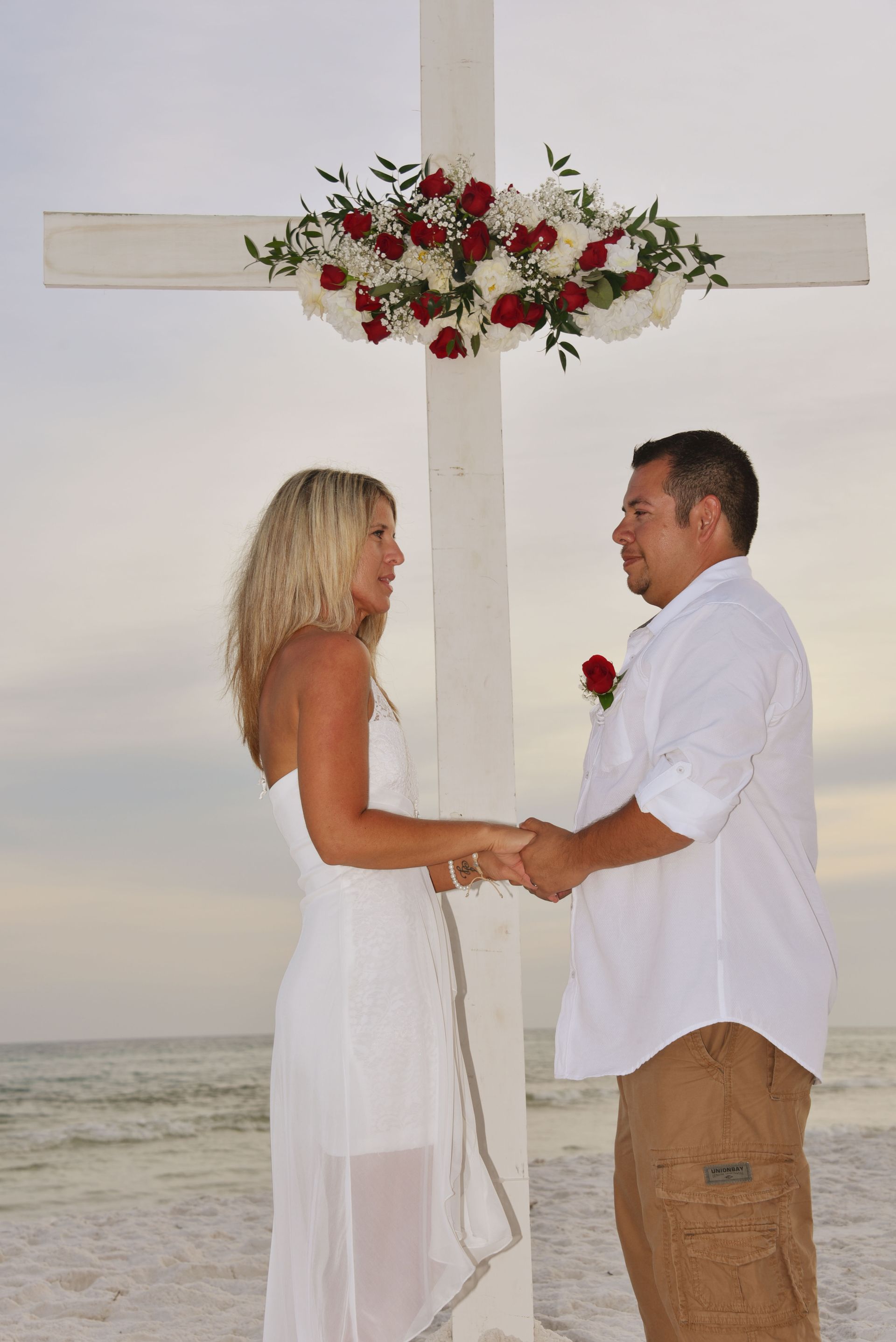 Couple exchanging vows on a beach under a flower-decorated cross at sunset