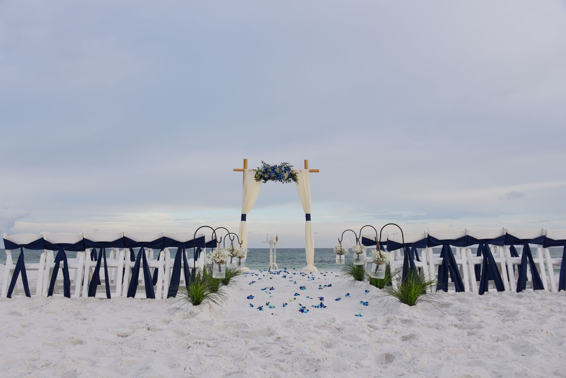 White wedding arch with blue ribbons on a snowy beach, surrounded by rows of dark chairs under an overcast sky