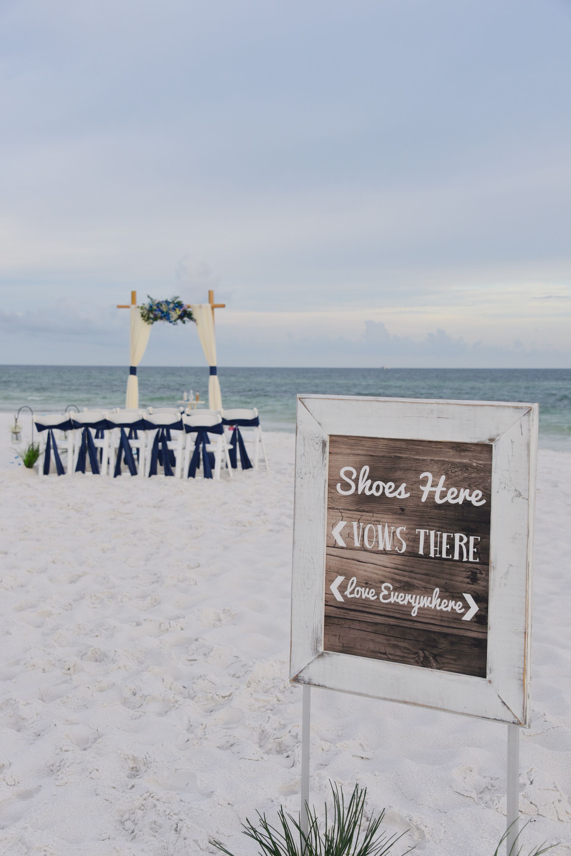 Beach wedding setup with white arch, blue chairs, and a sign on the sand reading “Share Here”
