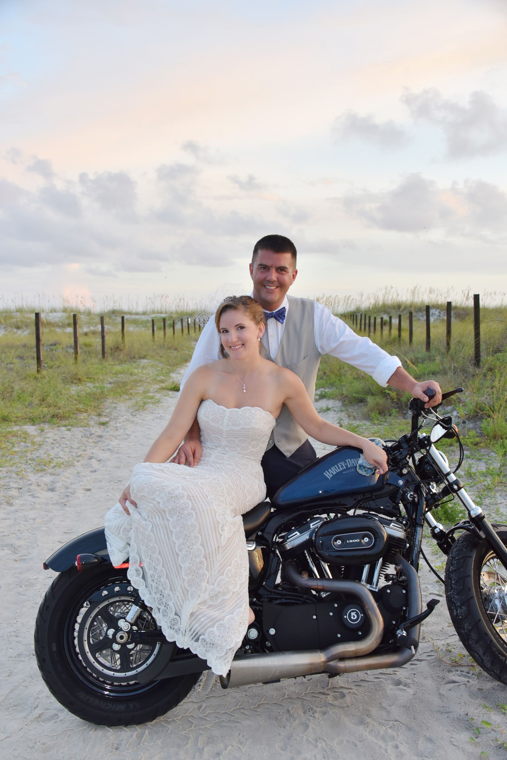 Couple in wedding attire on a black motorcycle on a gravel road at sunset