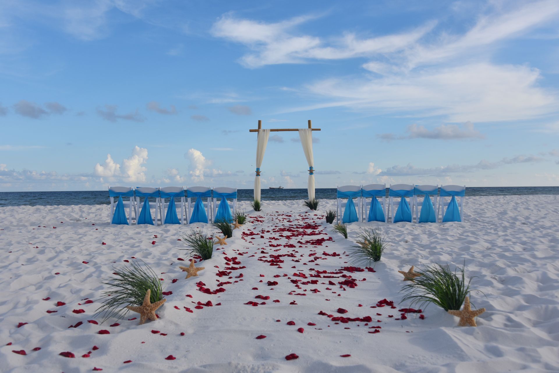 Beach wedding altar with white sand, blue chairs, scattered red petals, and a wooden arch under a cloudy sky