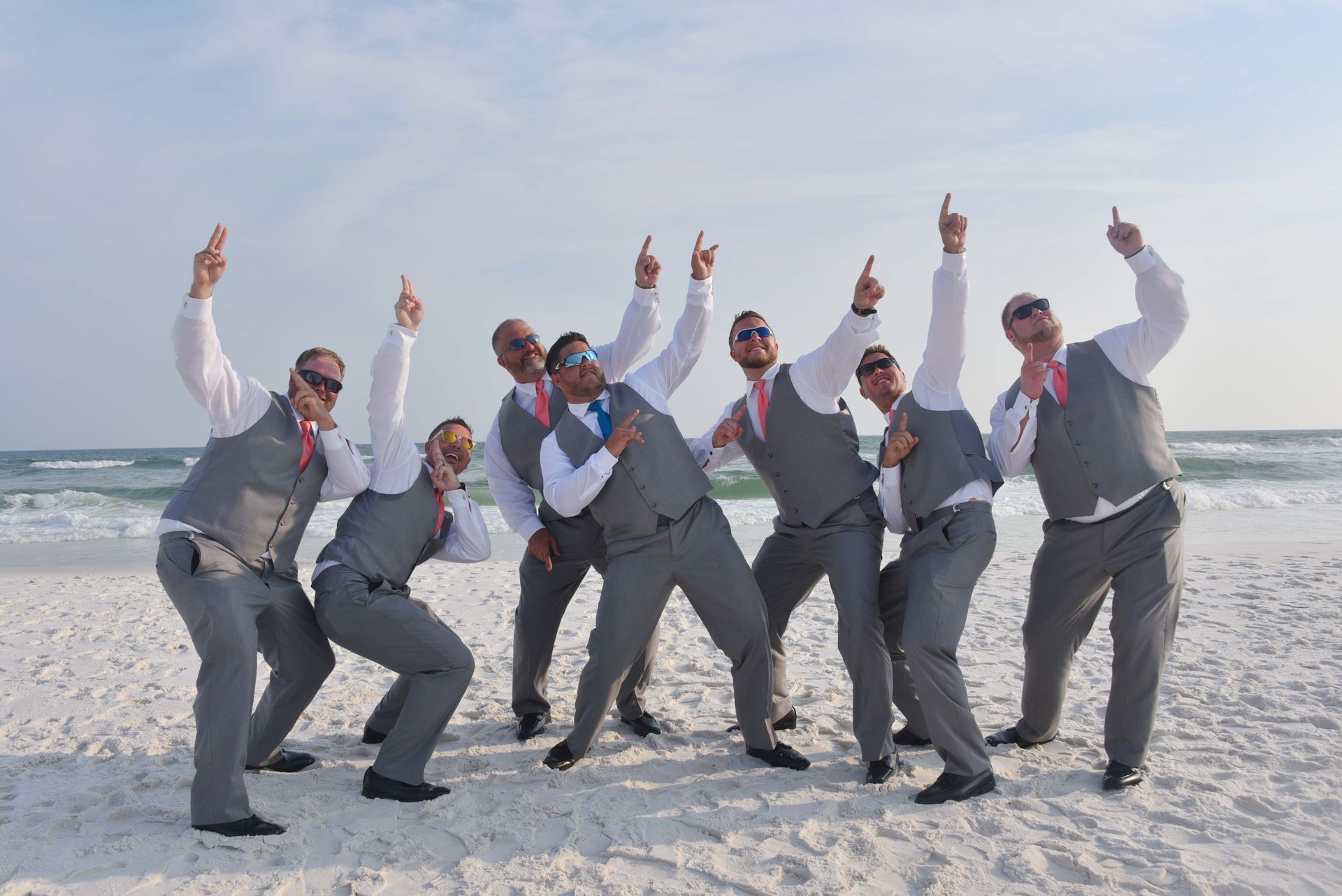Group of men in gray suits posing and pointing upward on a sunny beach by the ocean