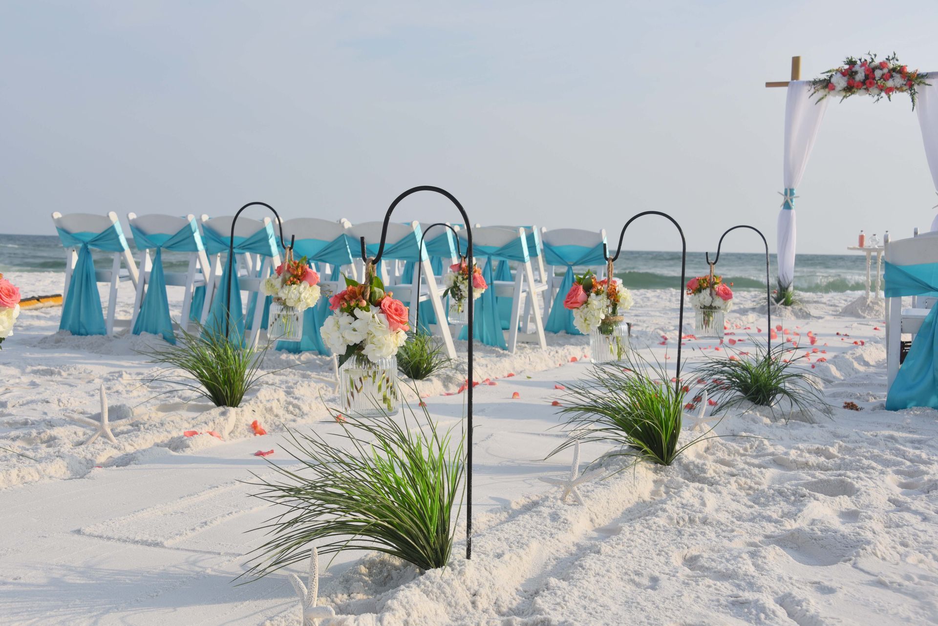 White sandy beach wedding setup with turquoise drapes, floral arches, and ocean in the background