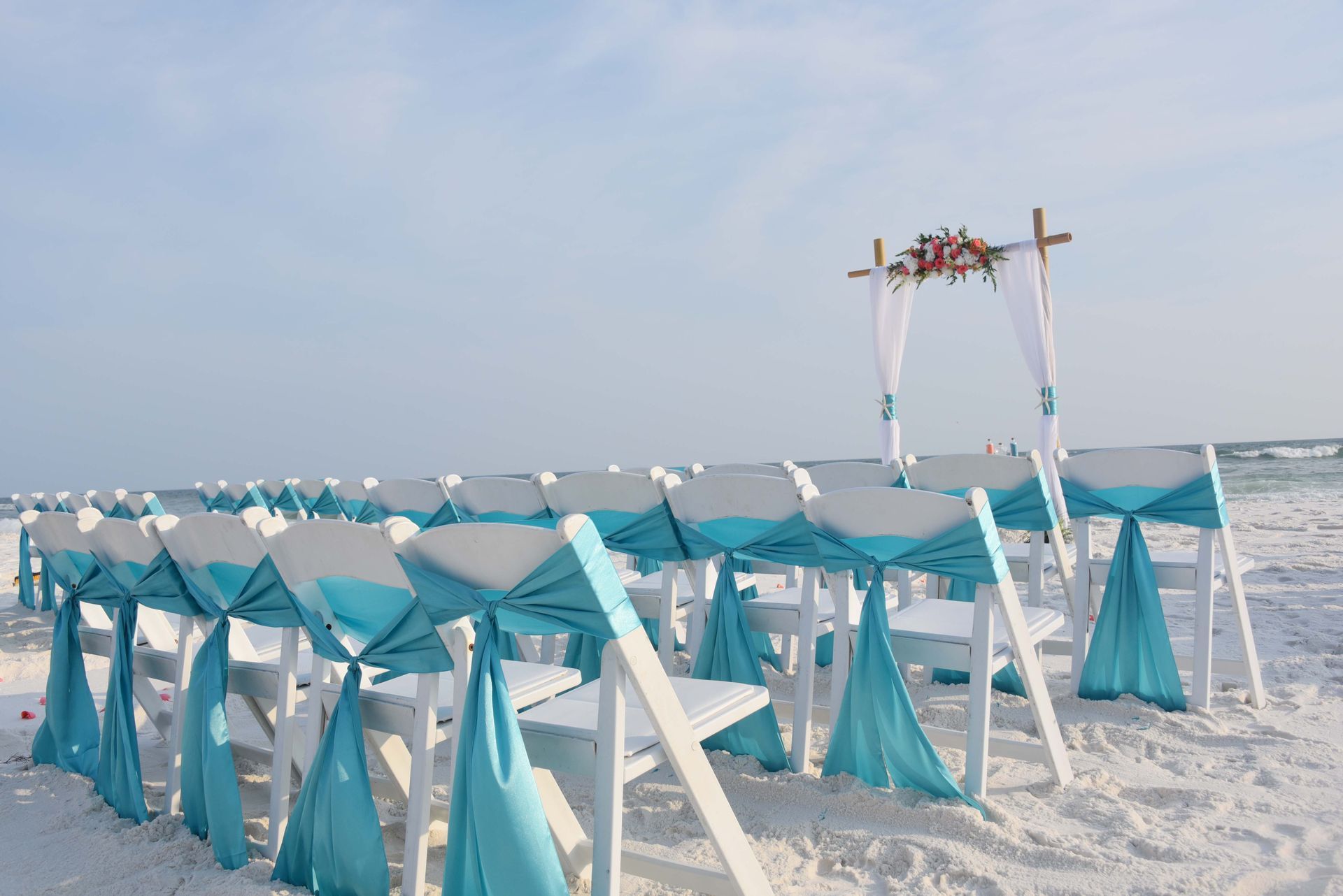 Beach wedding setup with turquoise-draped chairs and a floral arch on white sand by the sea