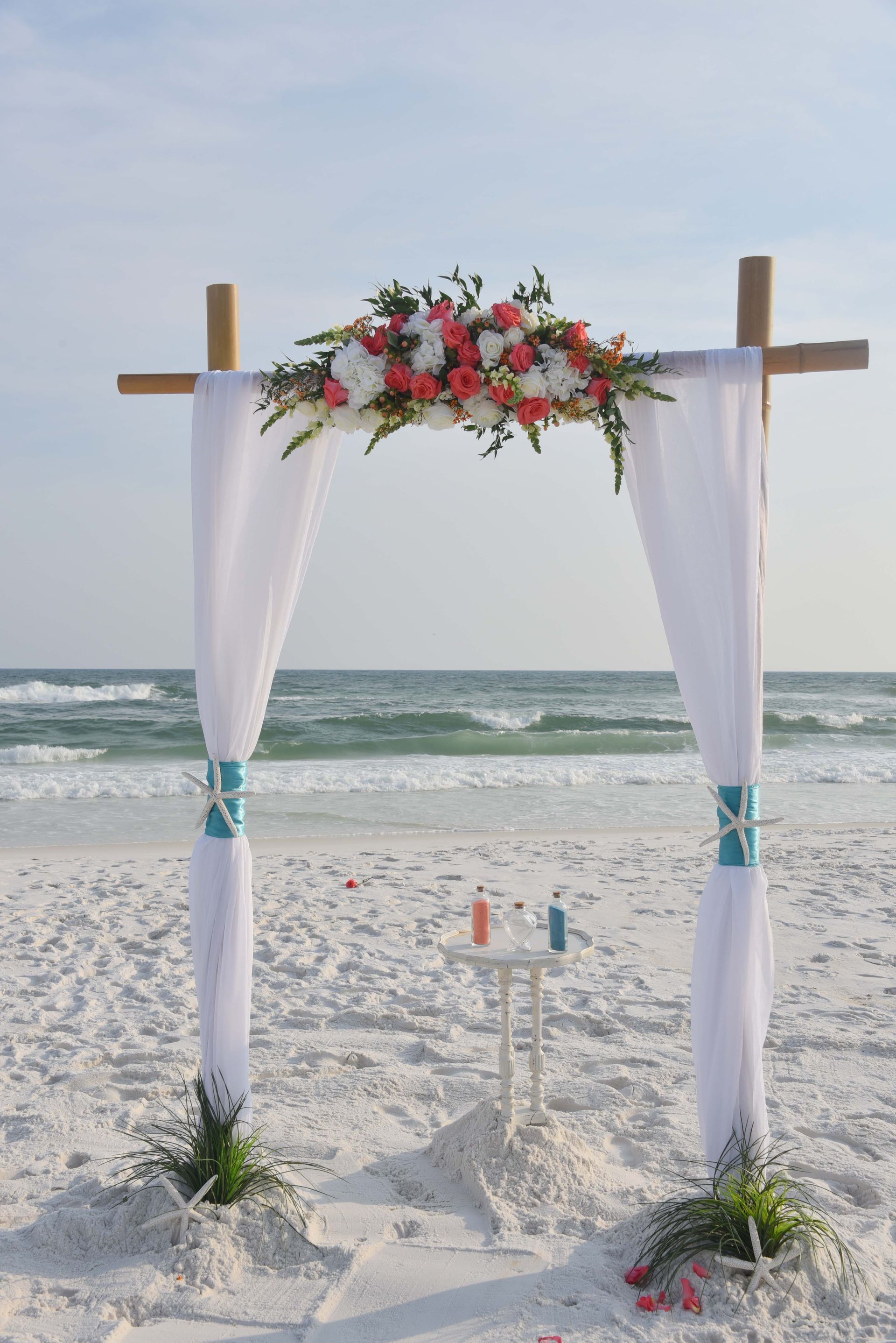 Beach wedding arch with white drapes and flowers on the shoreline at sunset