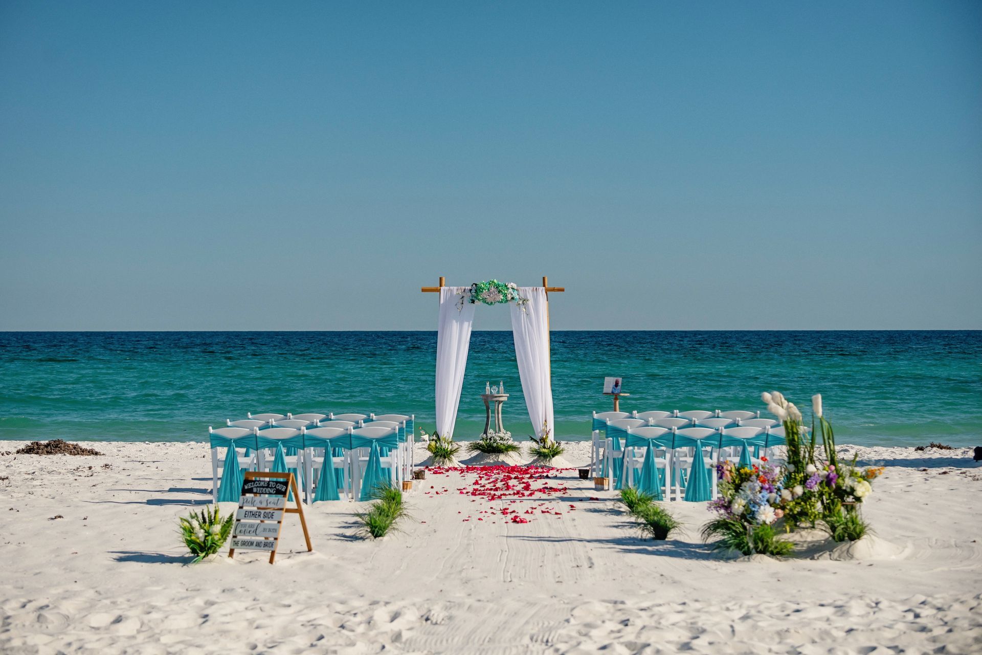 Beach wedding altar with white drapes, turquoise chairs, and floral aisle by the ocean