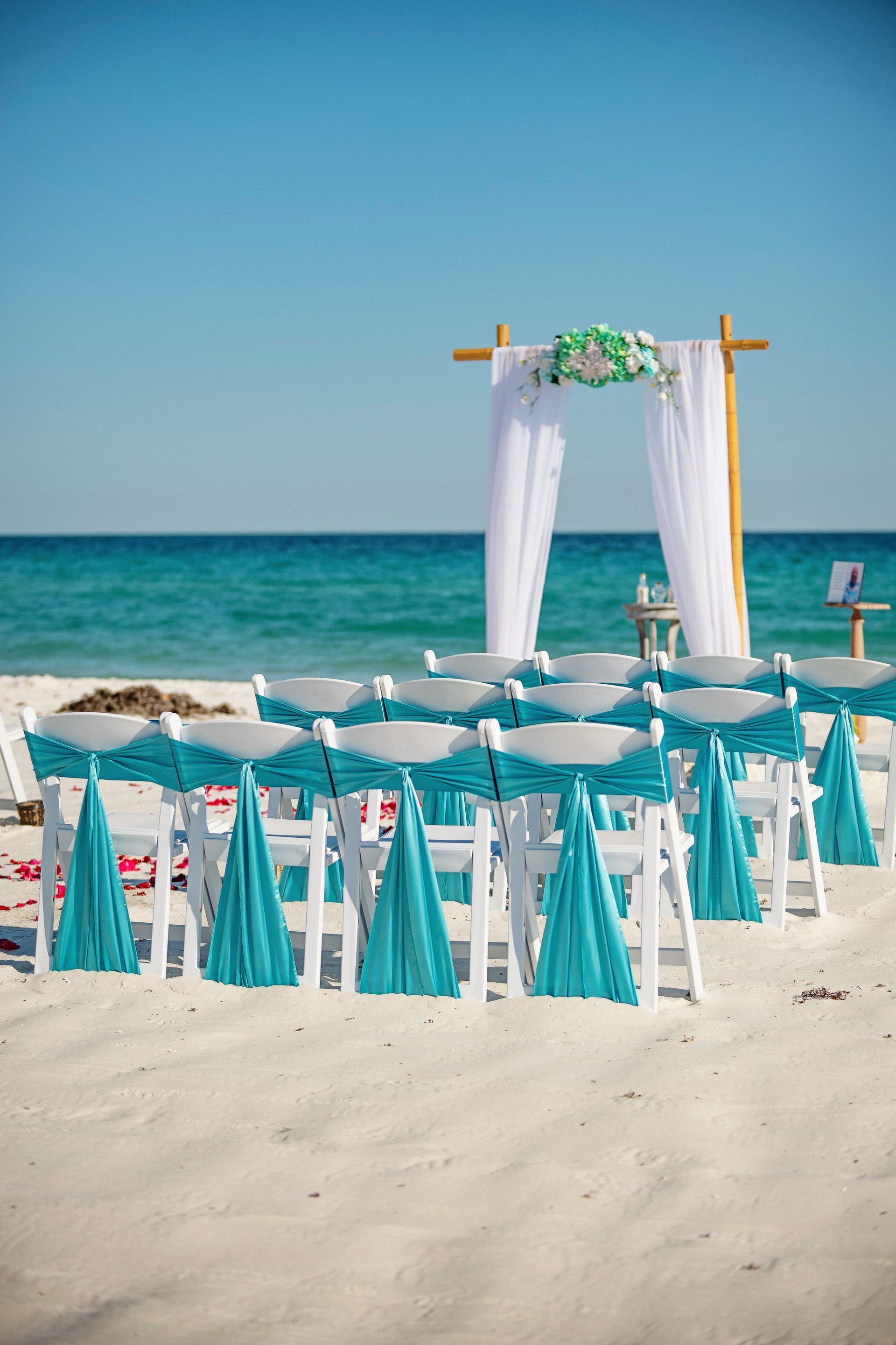 Beach wedding setup with white chairs, turquoise accents, and an arch by the ocean