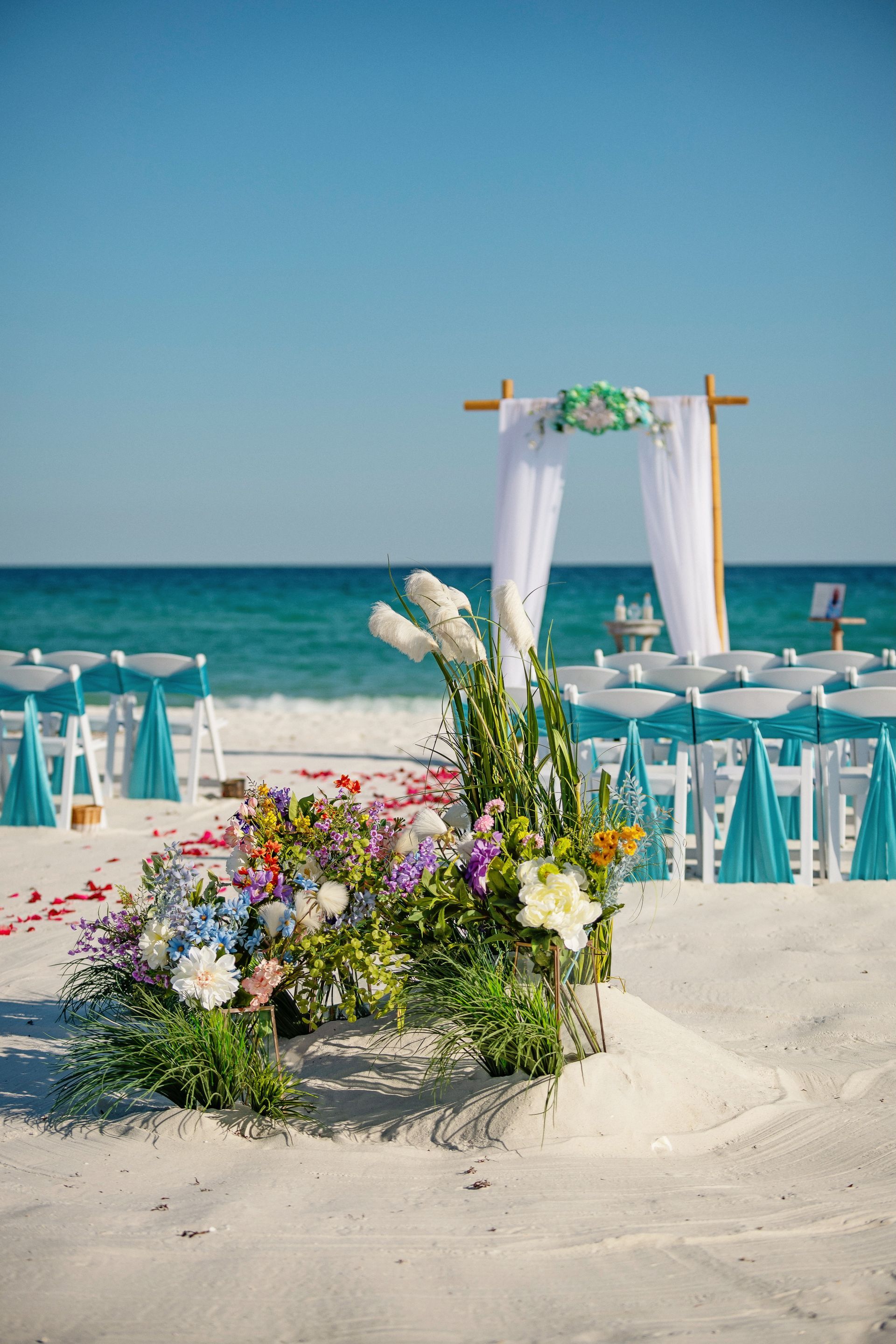 Beach wedding setup with white arch, teal chairs, and colorful flowers on the sand