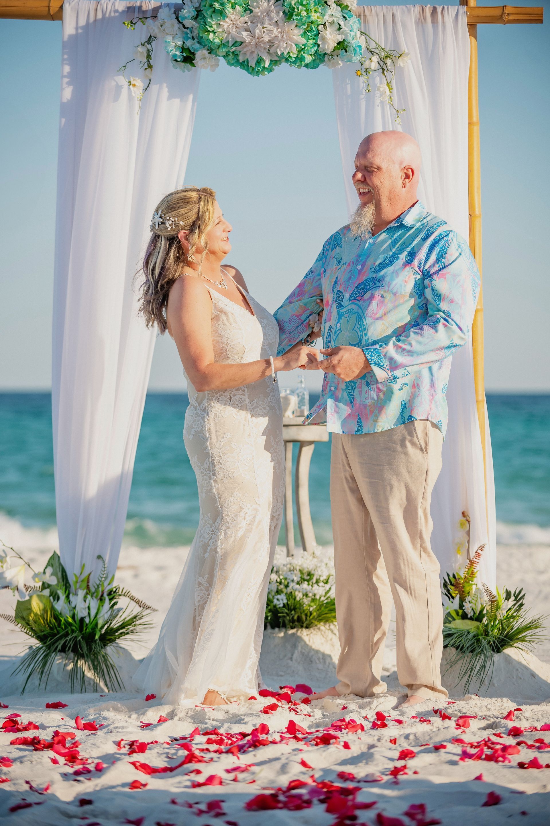 Beach wedding couple holding hands under a floral arch, standing on white sand with ocean behind them
