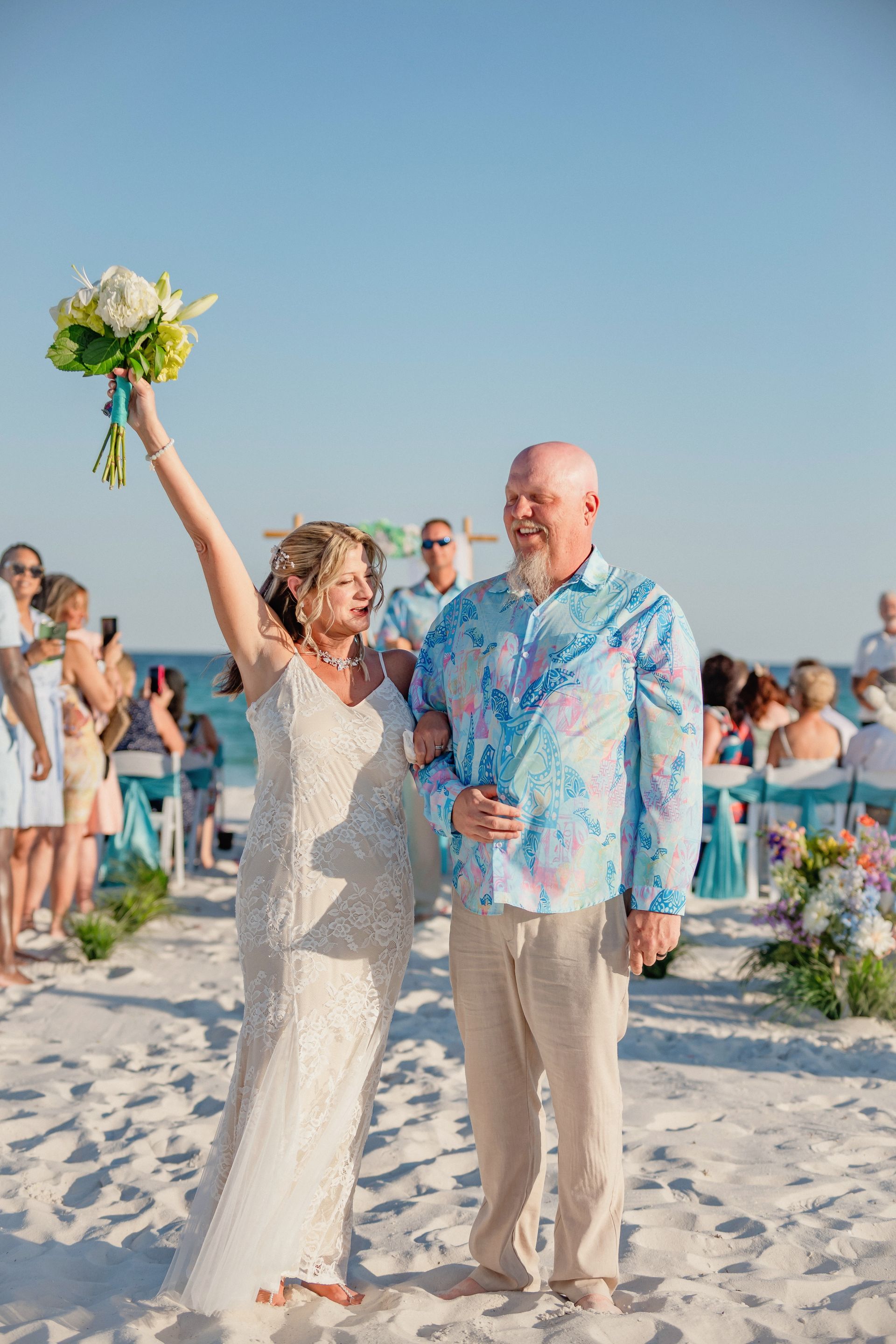 Bride raising bouquet on a sandy beach beside a smiling man, with wedding guests in the background