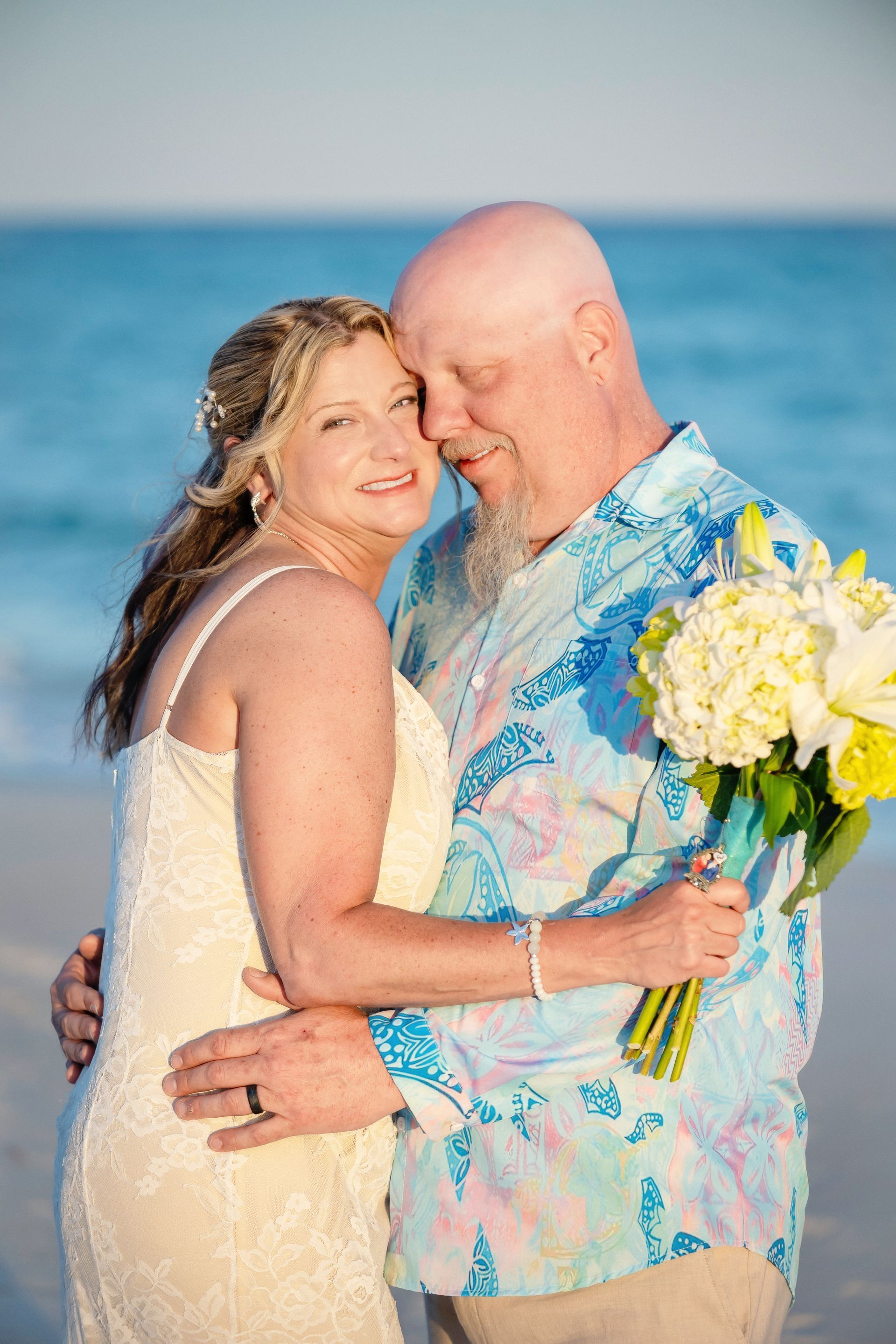 Couple embracing on a beach, smiling, with blue ocean and bouquet of white flowers