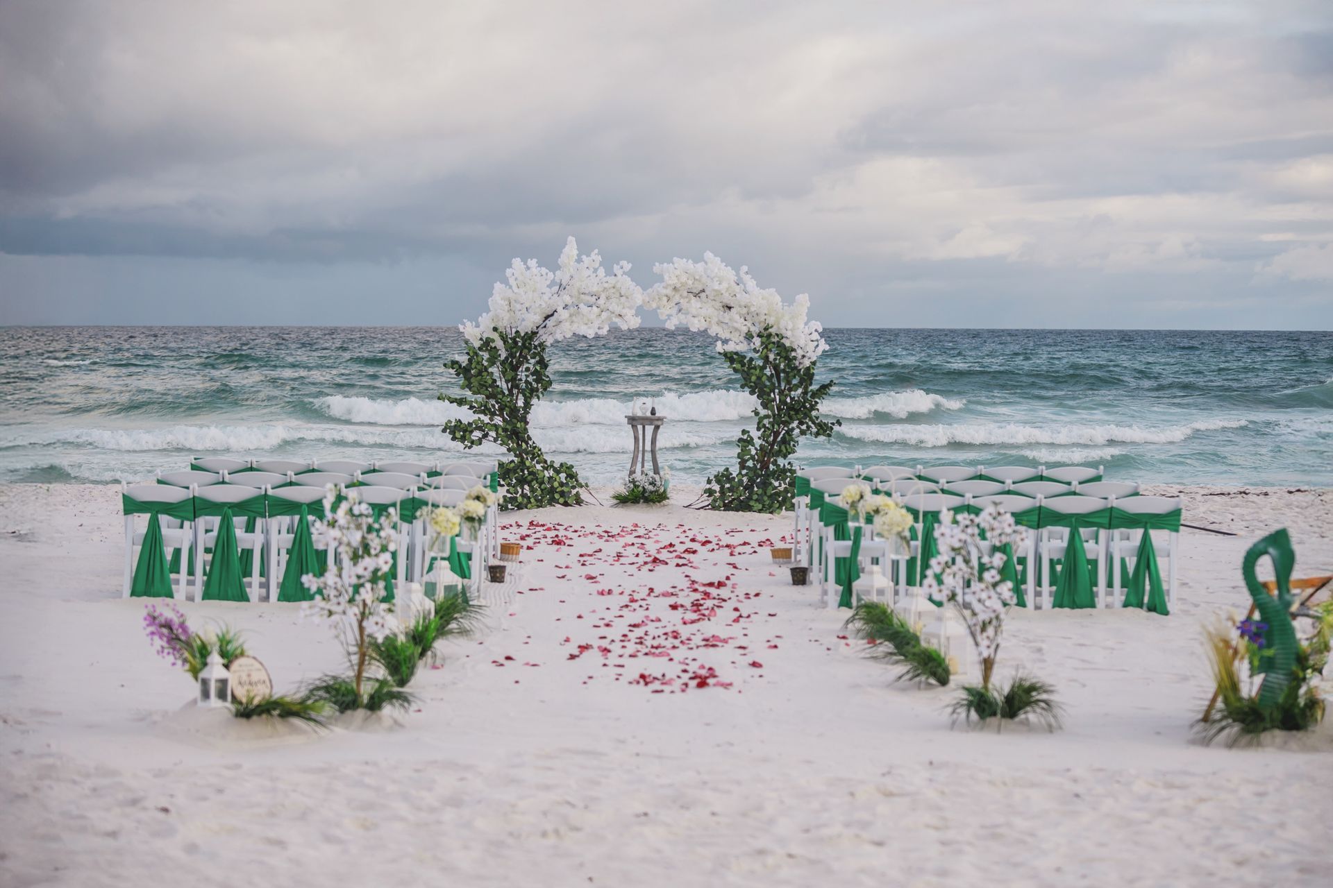 Beach wedding arch with white flowers, green chairs, and red petals on the sand by the ocean