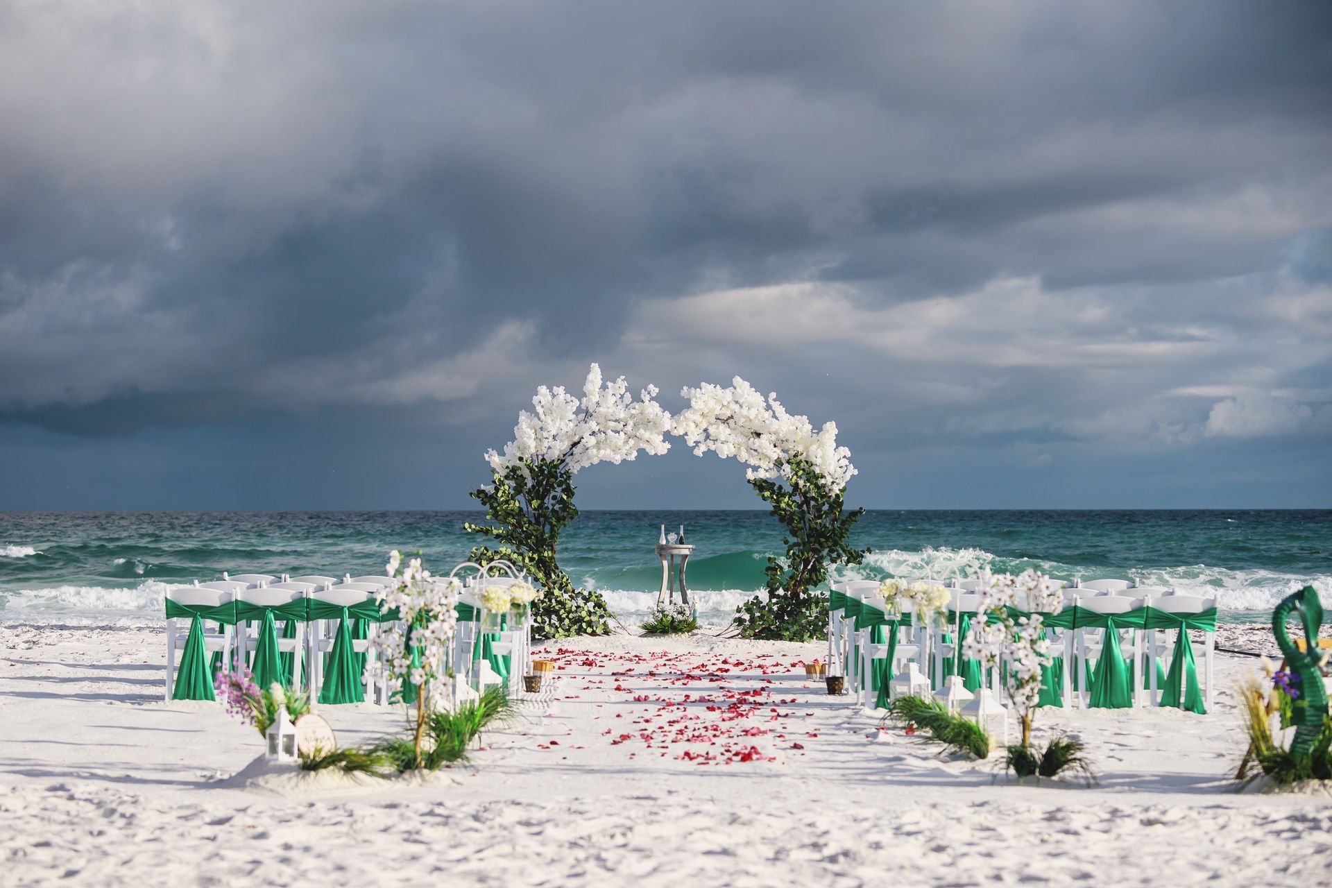 White floral arch on a decorated beach aisle with chairs, facing the ocean under cloudy skies.