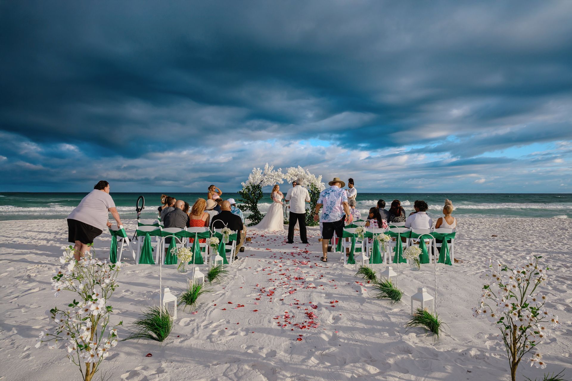 Beach wedding ceremony with white floral arch, green chairs, and guests under a cloudy sky