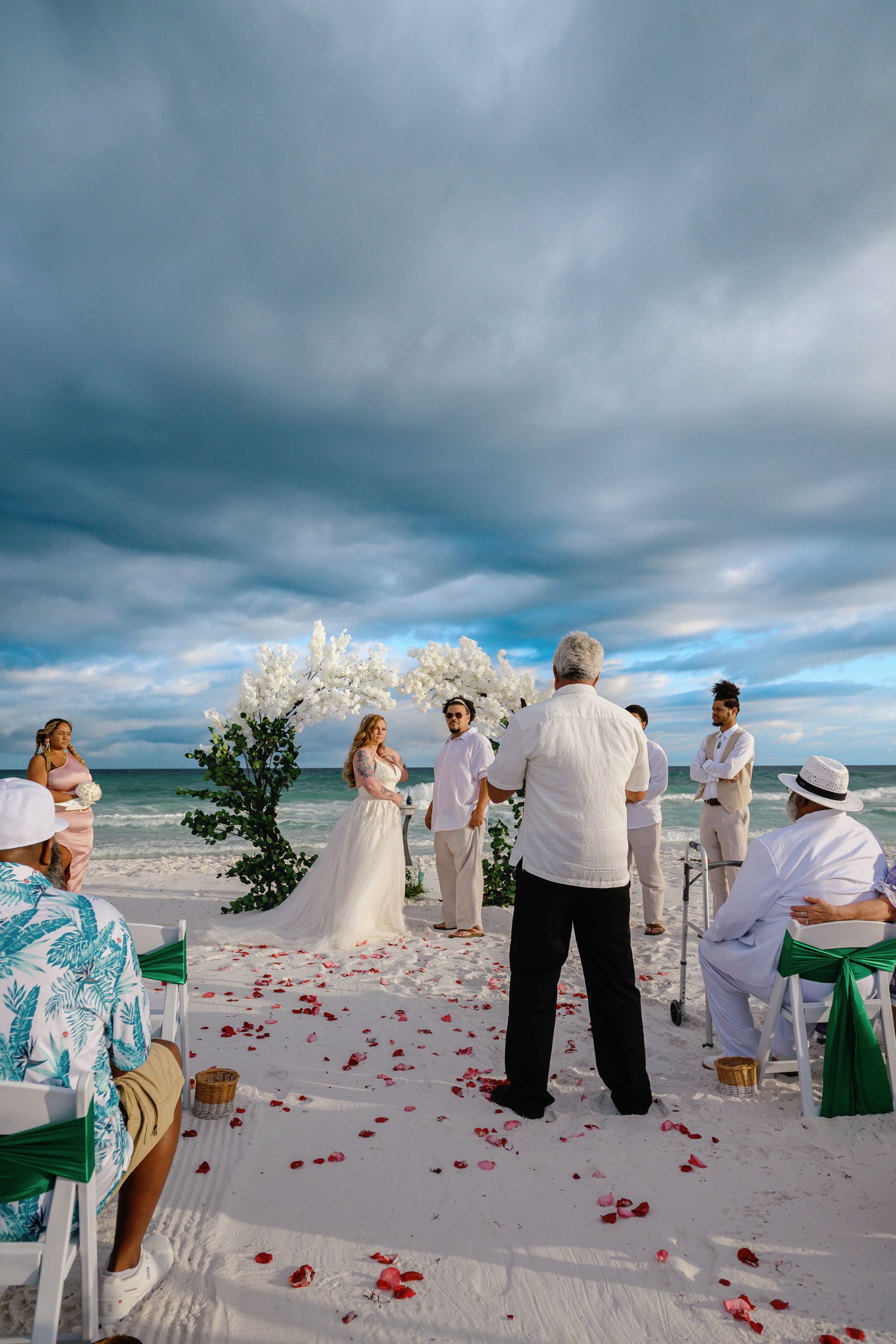 Beach wedding ceremony under cloudy sky with rose petals scattered along an aisle