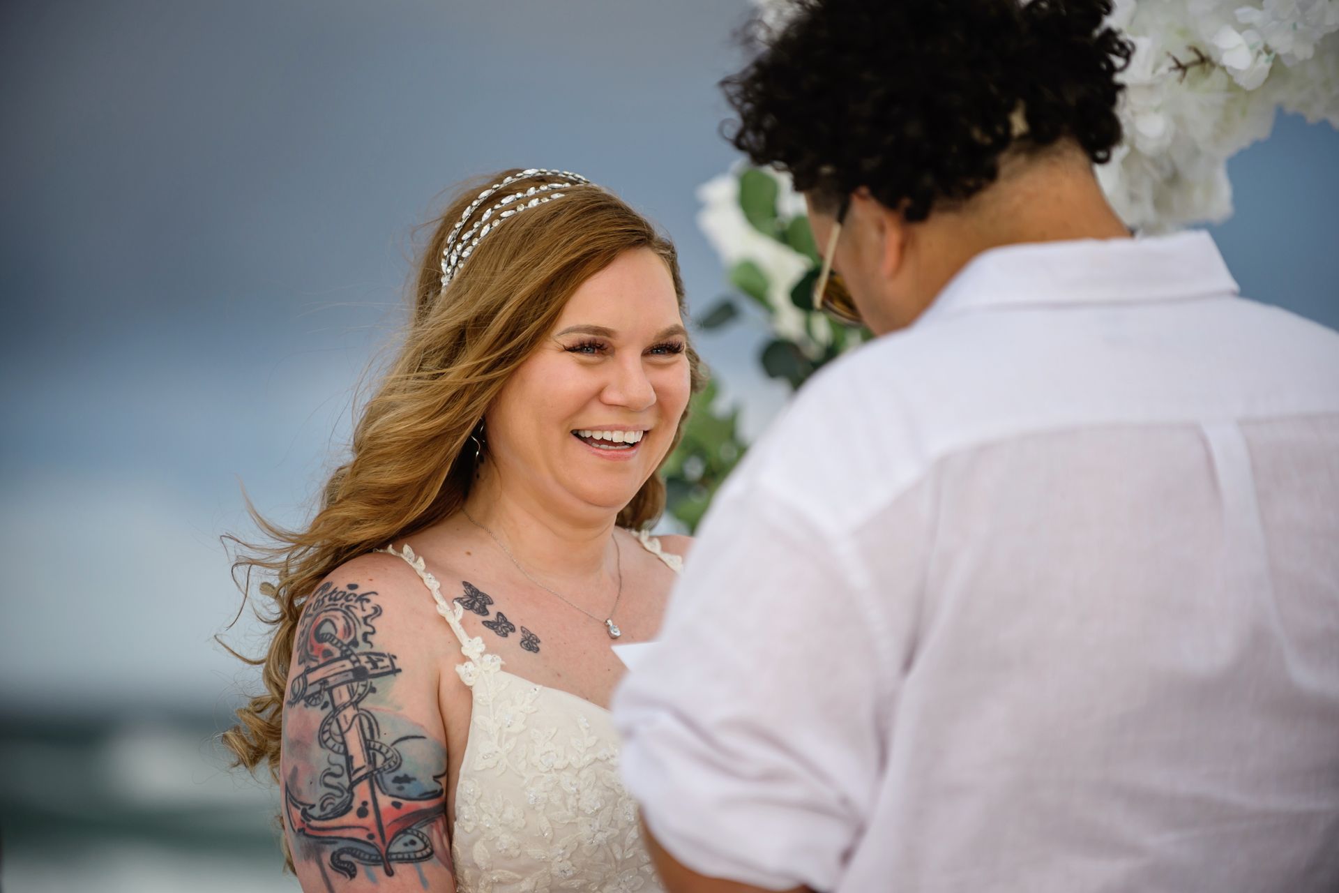 Smiling bride in a white dress with floral tattoo, facing a person in a white shirt outdoors