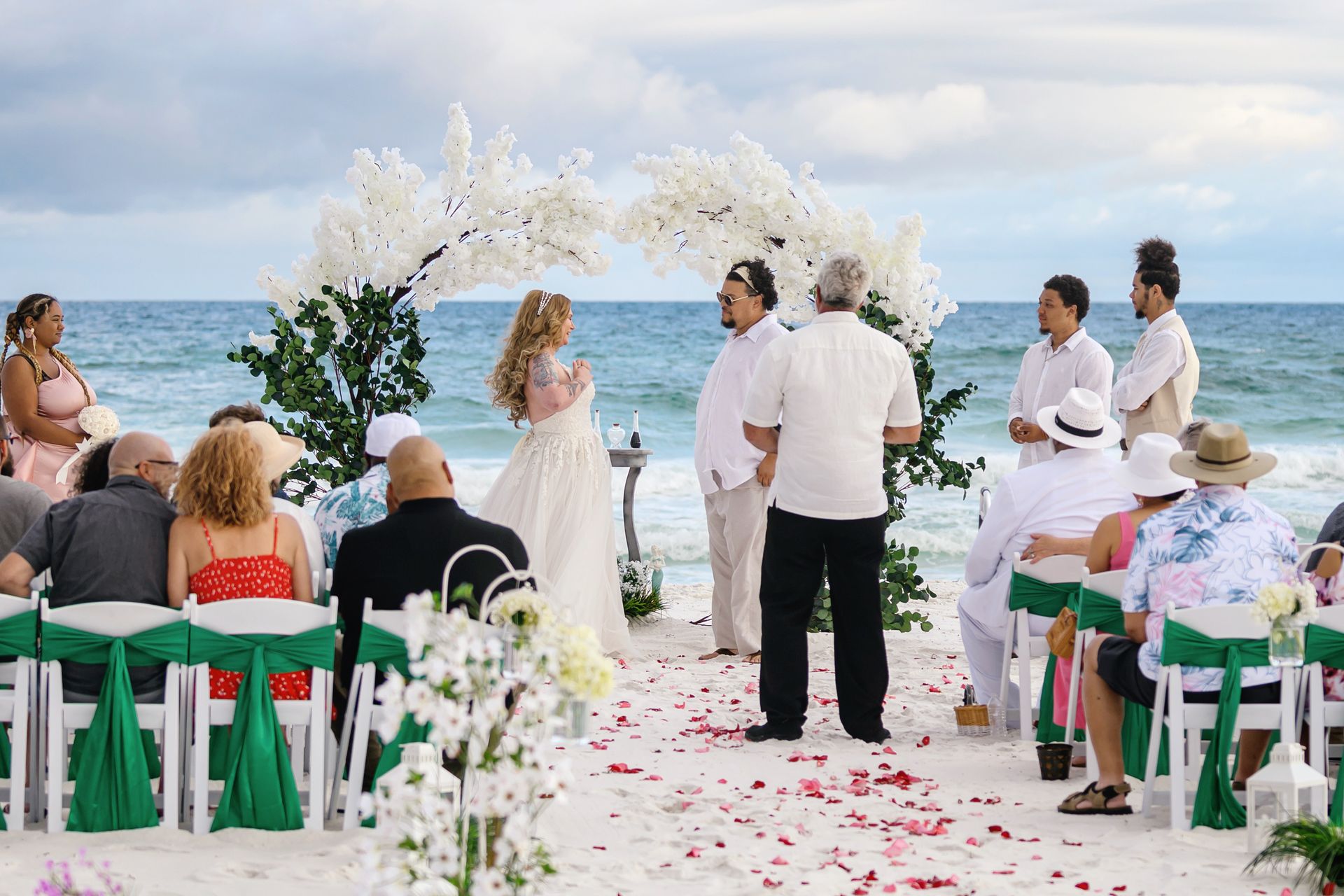 Beach wedding ceremony under a white floral arch, with seated guests and ocean in the background.