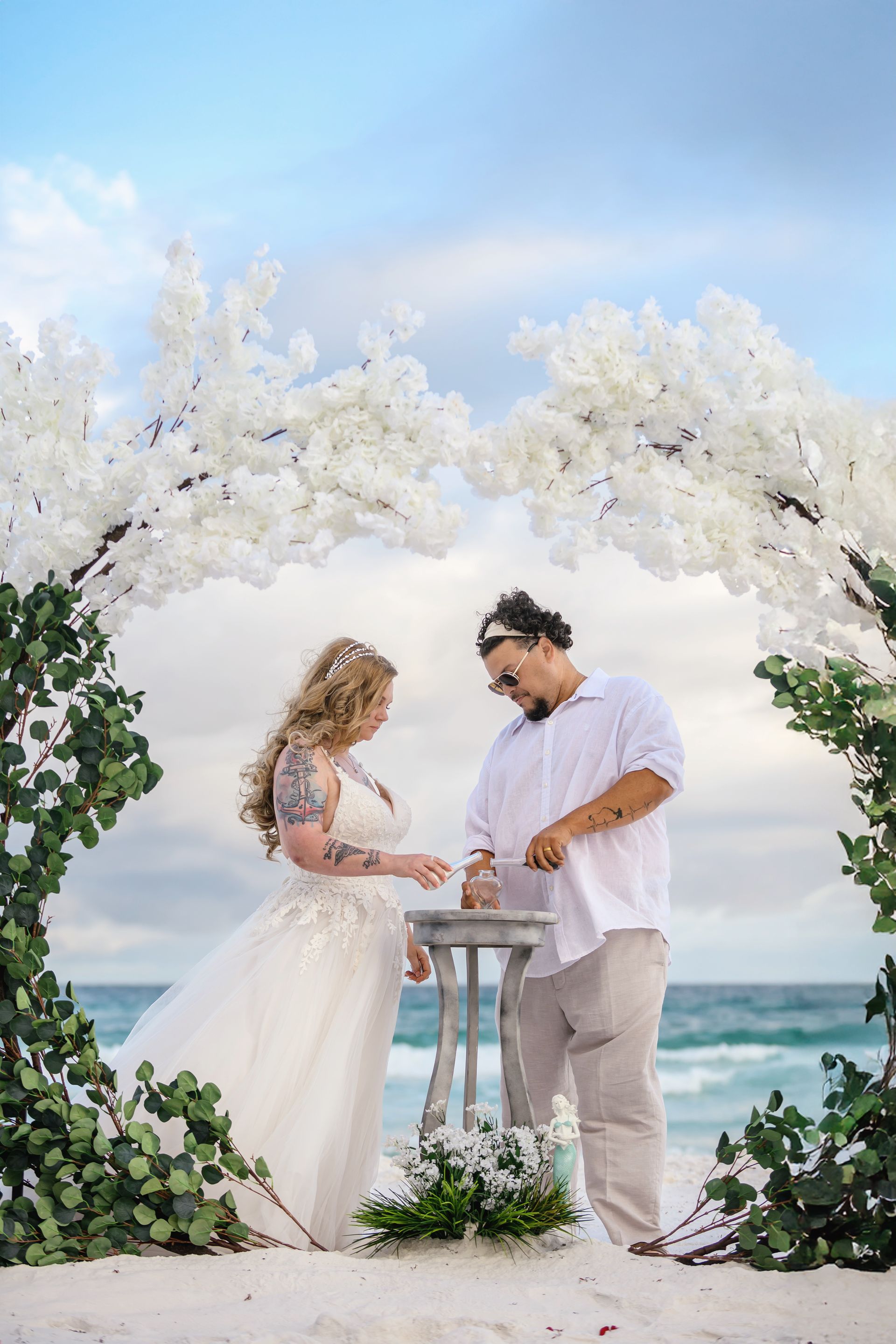 Couple exchanging rings under white floral arch by the ocean
