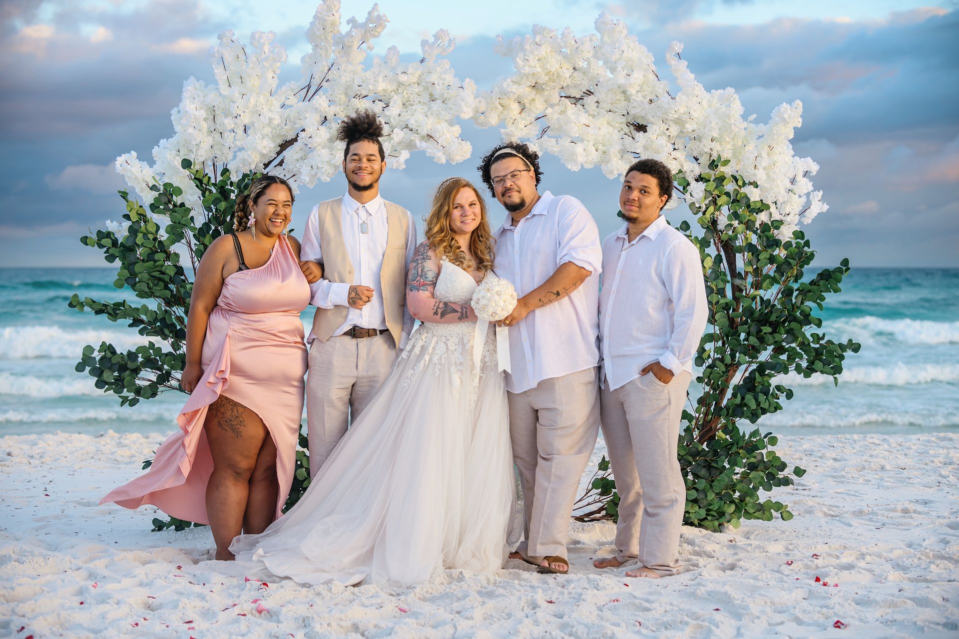 Five friends in light formalwear posing on a beach at sunset with white flowers and ocean waves