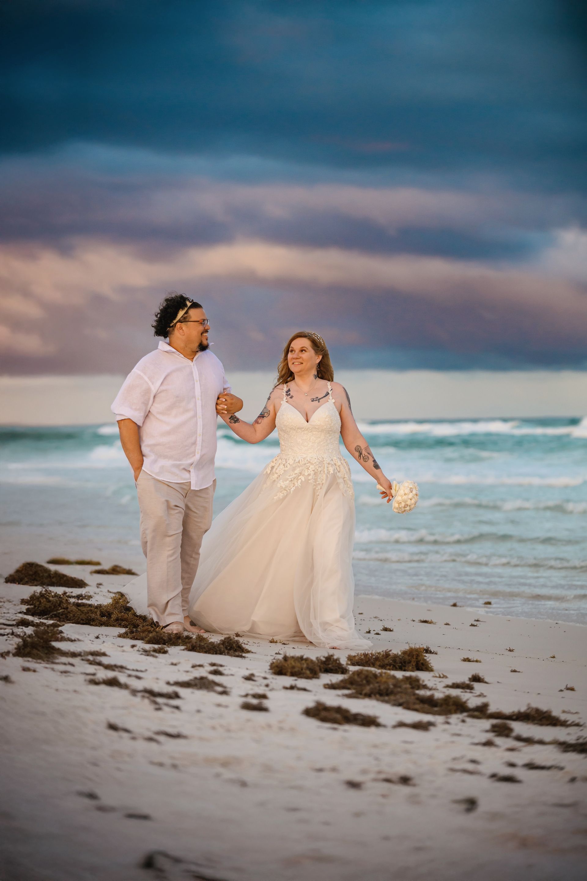 Couple in wedding attire walking hand in hand on a beach at sunset.