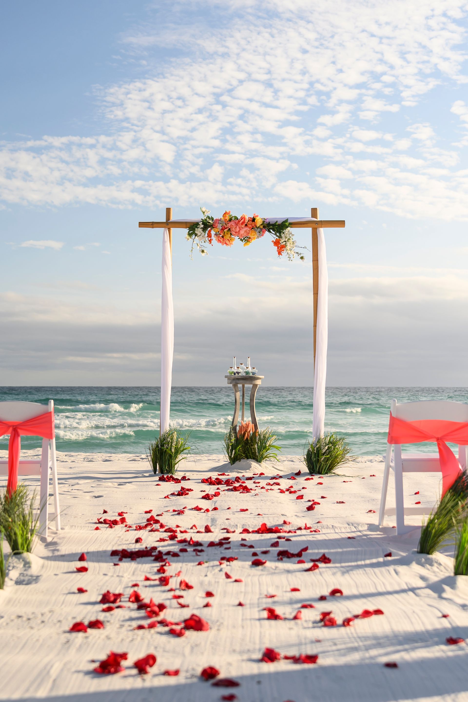 Beach wedding arch with white drapes, red petals, and ocean view at sunset