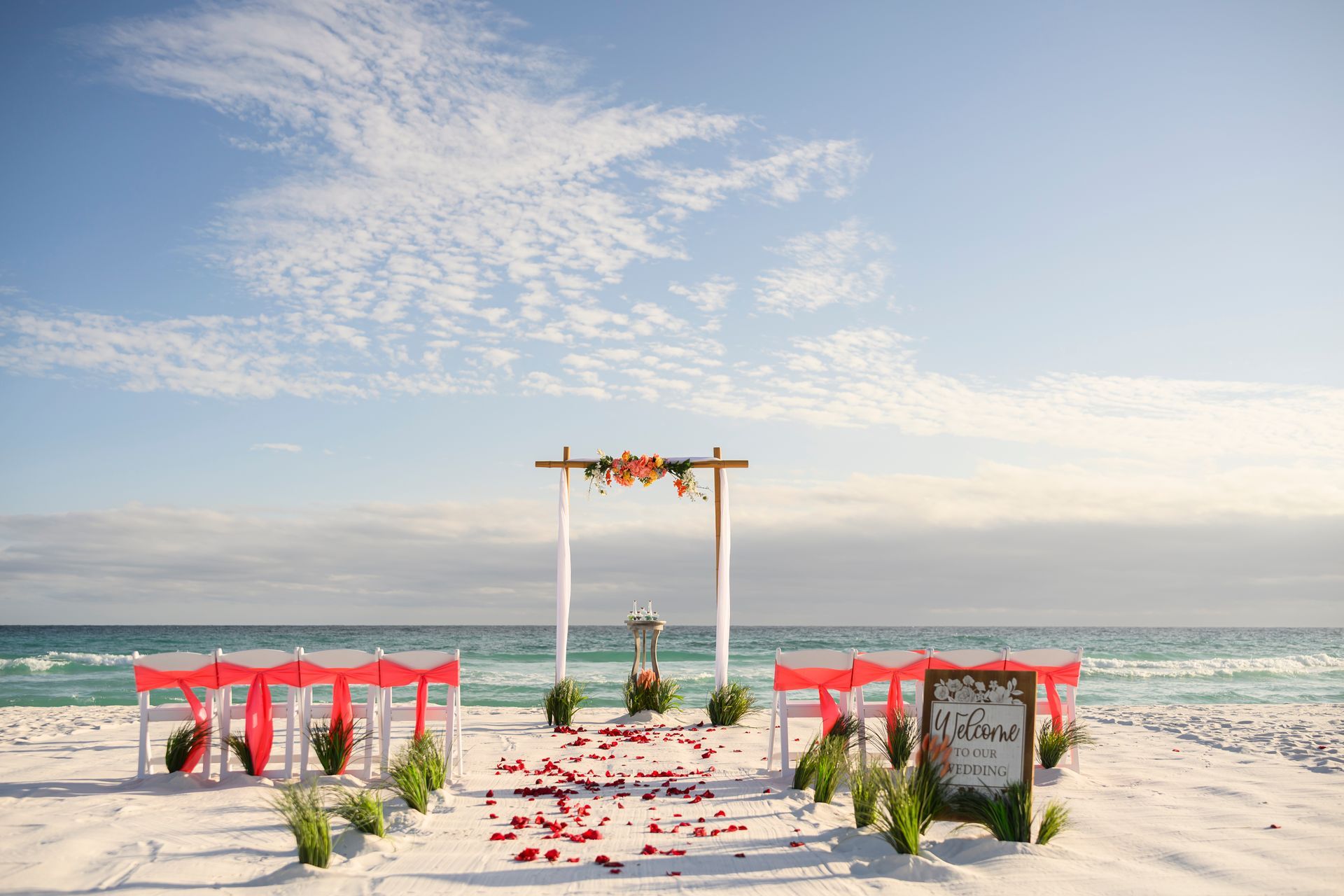 Beach wedding setup with white arch, red drapes, flower petals, and ocean backdrop