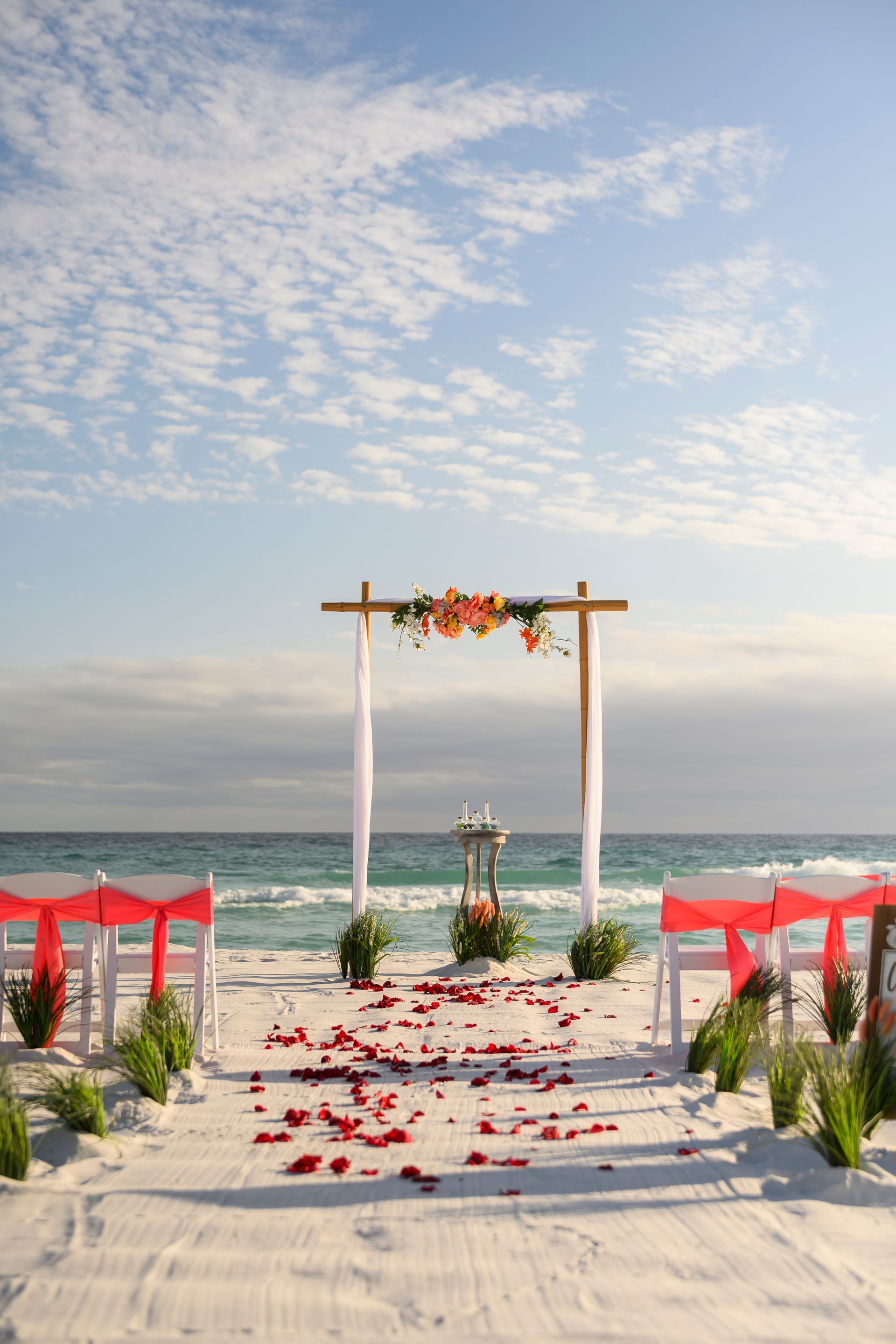 Beach wedding arch on a flower-strewn aisle, flanked by red chairs, with the ocean behind.