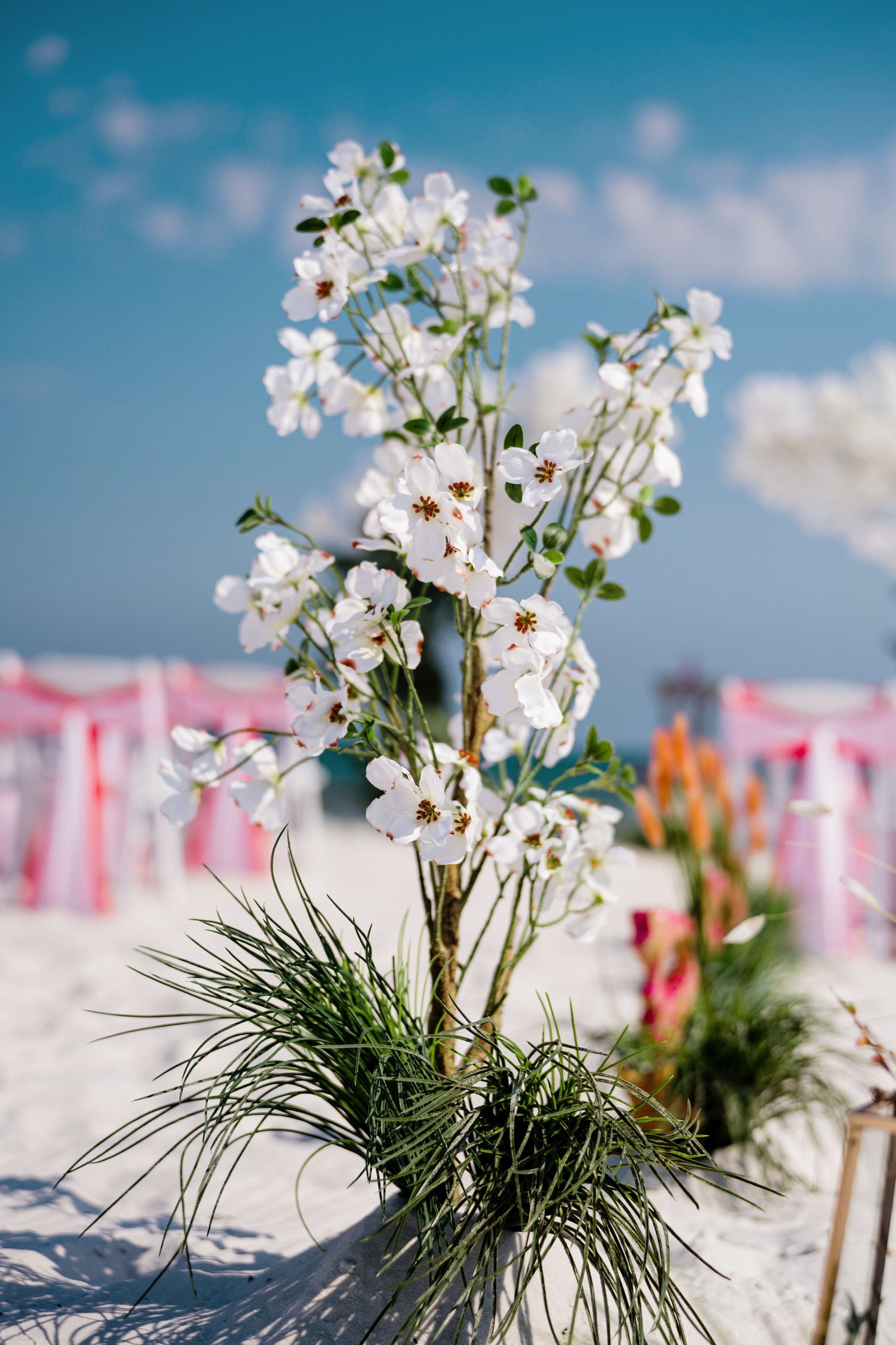White floral centerpiece in a glass vase on a beach wedding table with pink chairs and blue sky