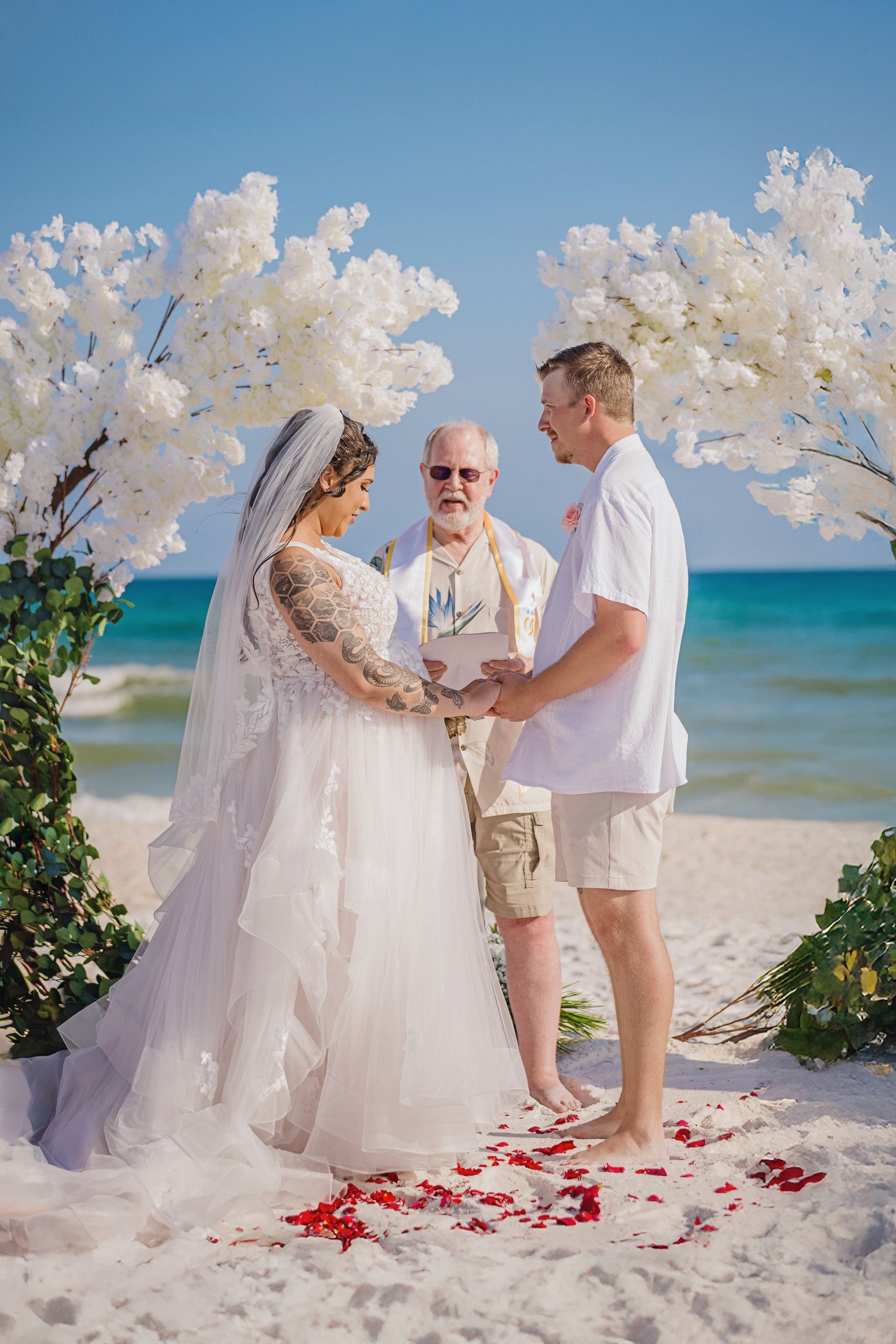 Beach wedding ceremony under white floral arches, with officiant and couple holding hands by the ocean.