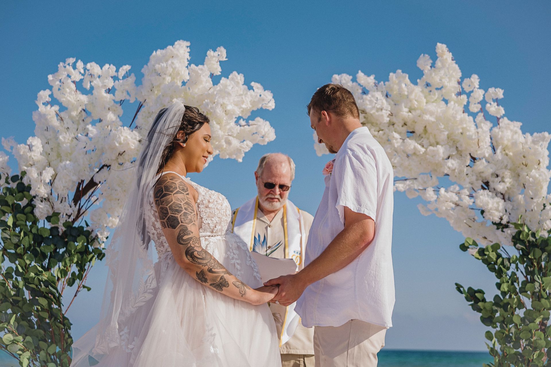 Bride and groom holding hands at a beach wedding arch with white flowers and an officiant behind them