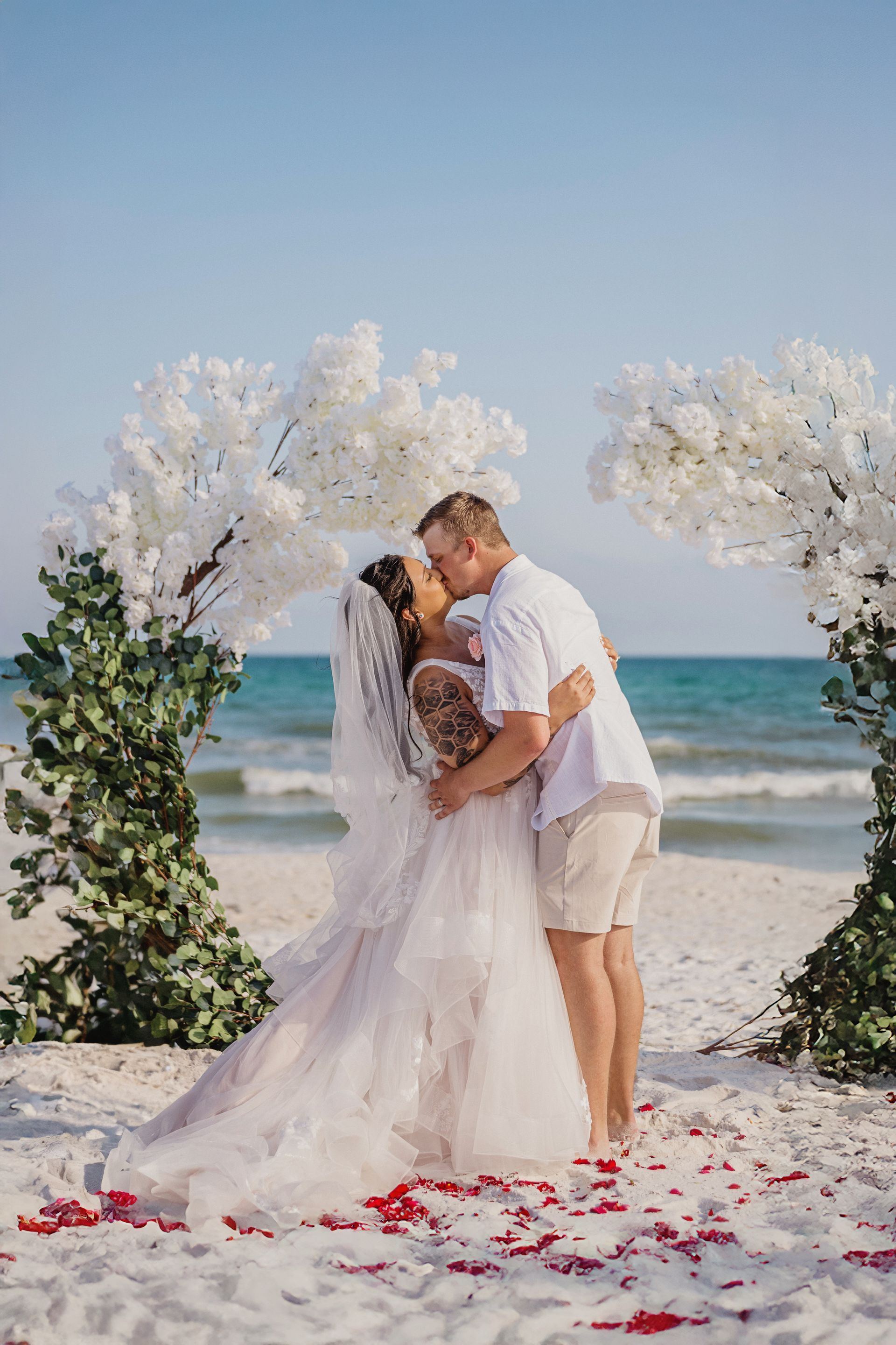 Couple kissing on a flower-lined beach wedding arch by the ocean