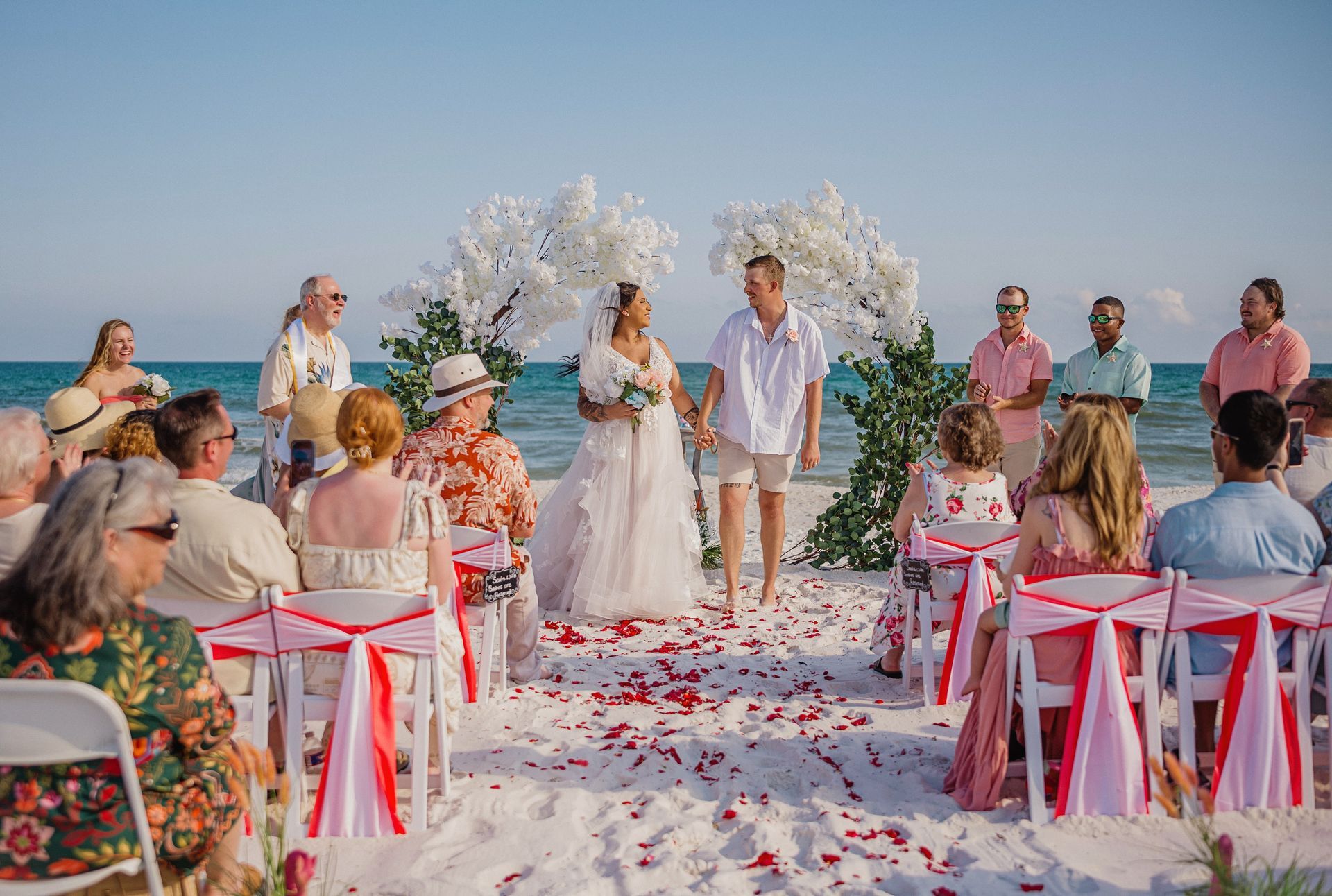 Beach wedding ceremony with bride and groom under floral arch, guests seated on chairs with pink ribbons
