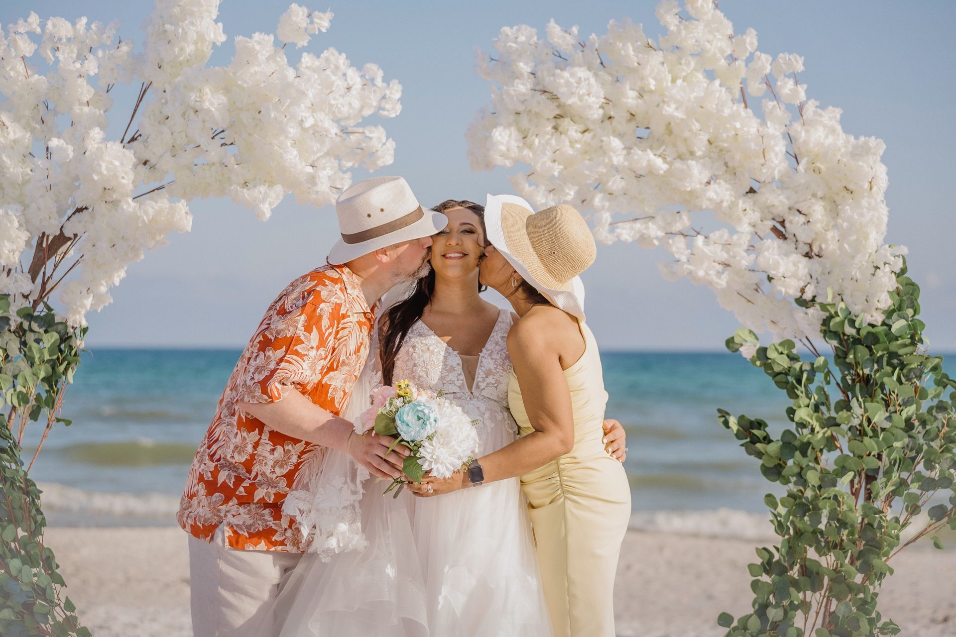 Three people in white and orange outfits celebrate a beach wedding under white floral arches.